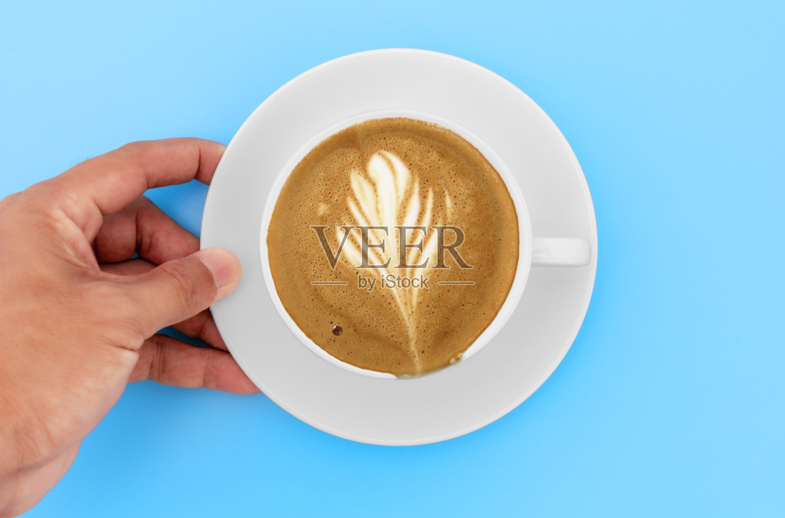 俯视图手握白色咖啡杯与卡布奇诺咖啡和奶油形状在白色杯子。在咖啡店的蓝色桌子上，手持咖啡杯的男人。照片摄影图片