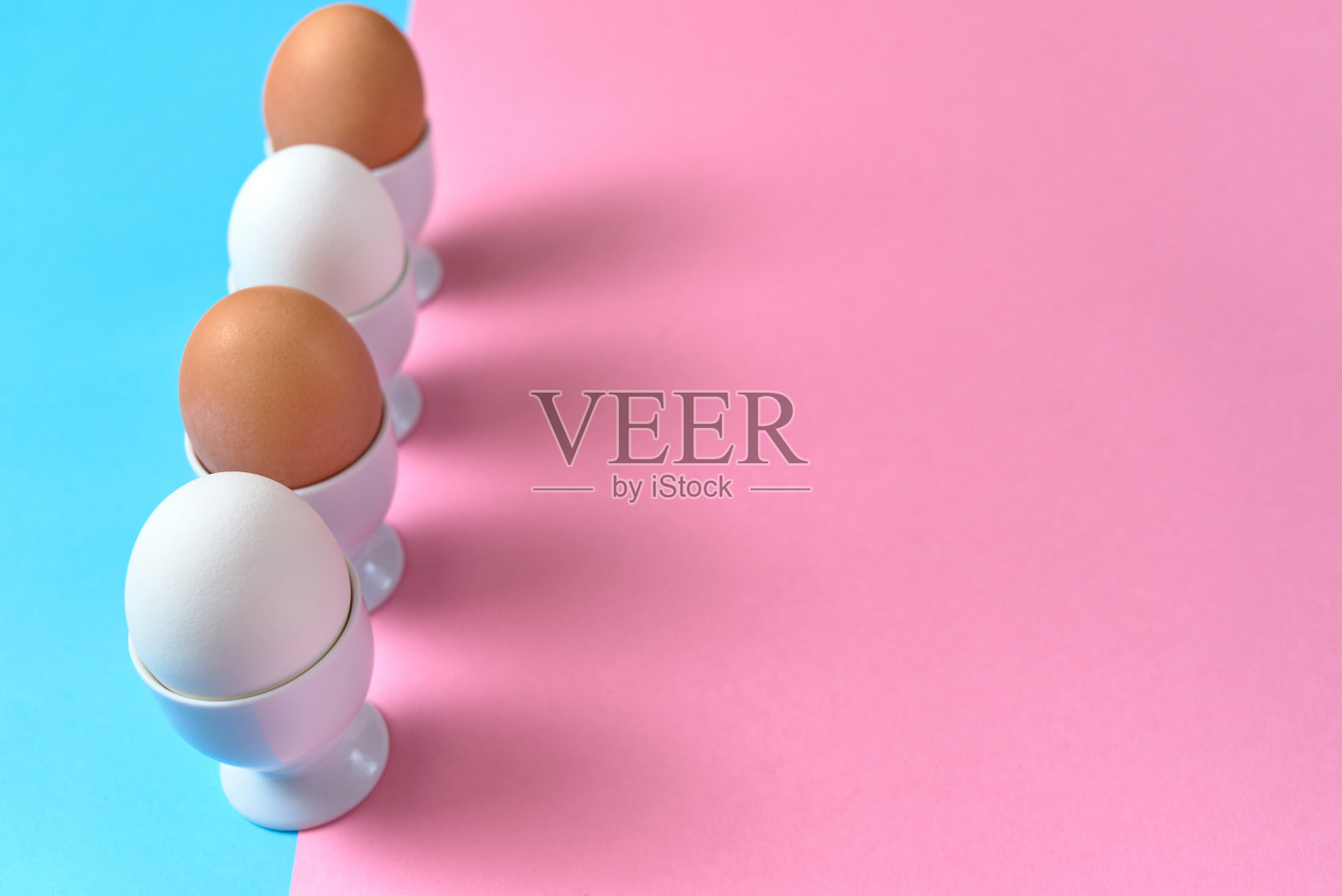 蛋站在蛋杯上，蓝色和粉色的粉彩背景，复制空间。煮熟的鸡蛋在纸底色上有两种色调。健康食品的概念。复活节彩蛋照片摄影图片