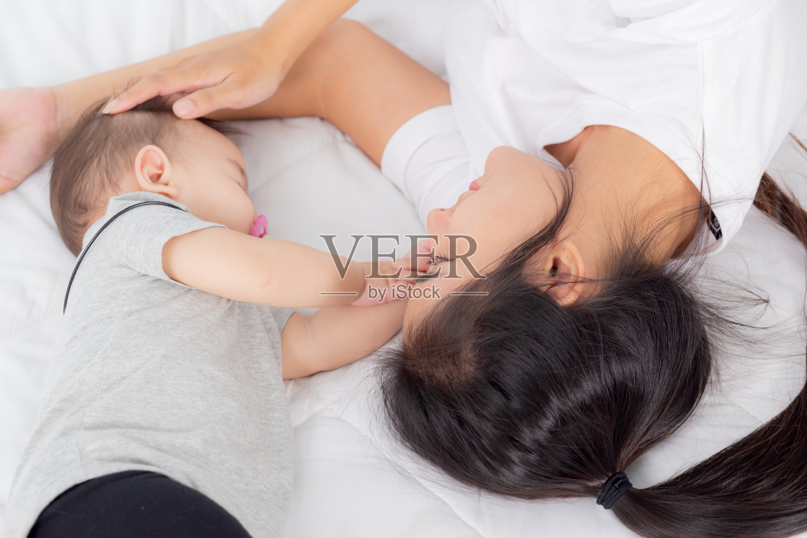 年轻的亚洲母亲在卧室的床上温柔地抚摸着小女婴的头，妈妈对新生儿的爱与呵护，女人与孩子一起表达着父母与女儿、家庭观念。照片摄影图片