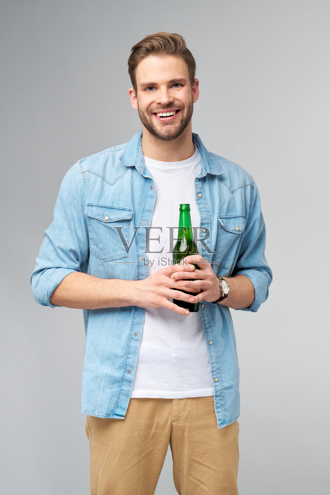 一个年轻人穿着牛仔裤衬衫，拿着一瓶啤酒站在灰色背景照片摄影图片