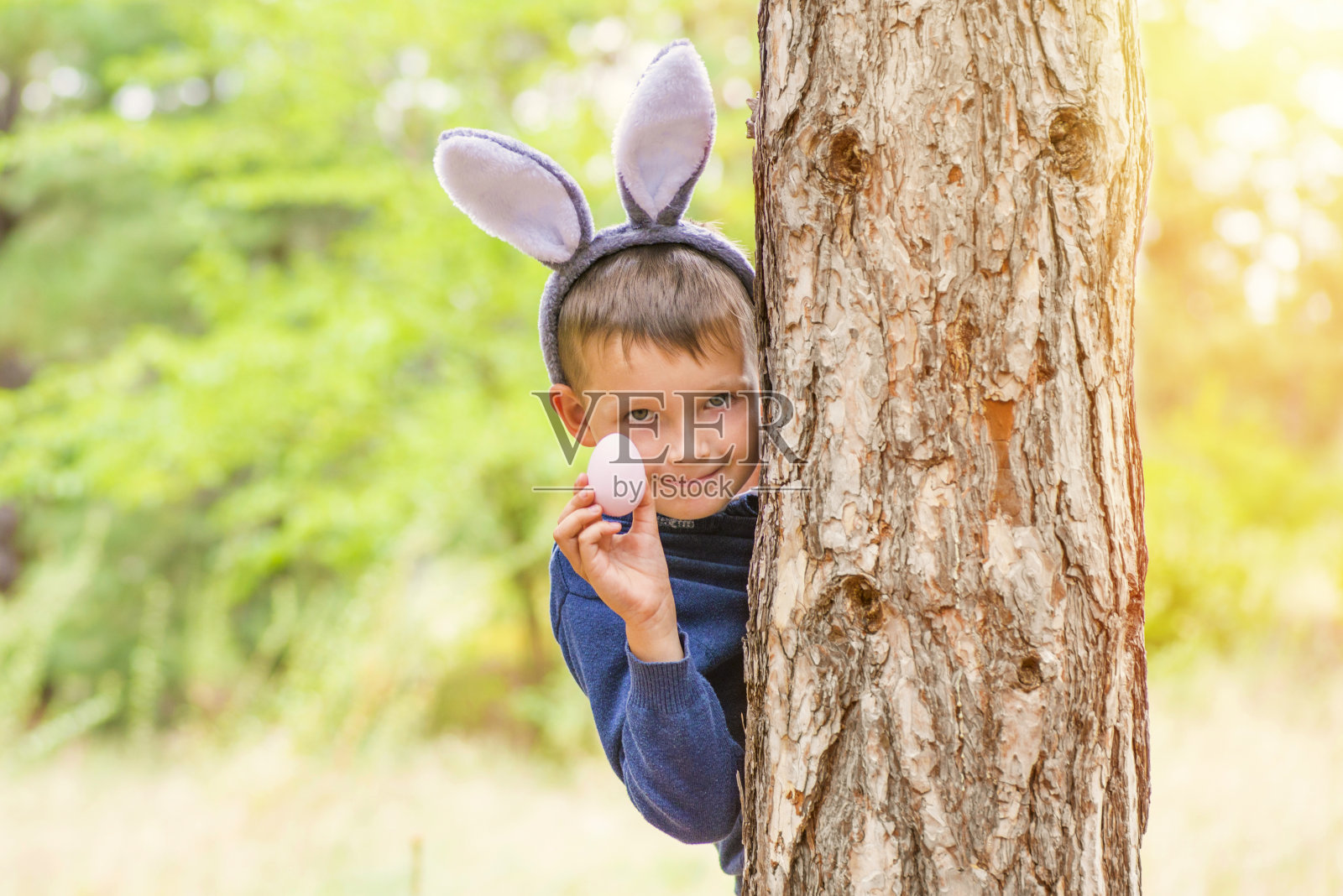 一个长着兔子耳朵的可爱小男孩站在树后，手里拿着复活节彩蛋。复活节快乐。照片摄影图片