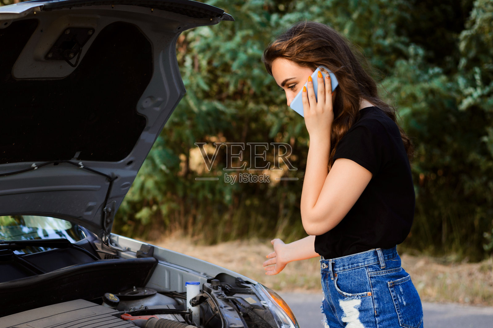 女司机打电话给汽车维修机构，描述了汽车的问题，看了看汽车打开的引擎盖照片摄影图片