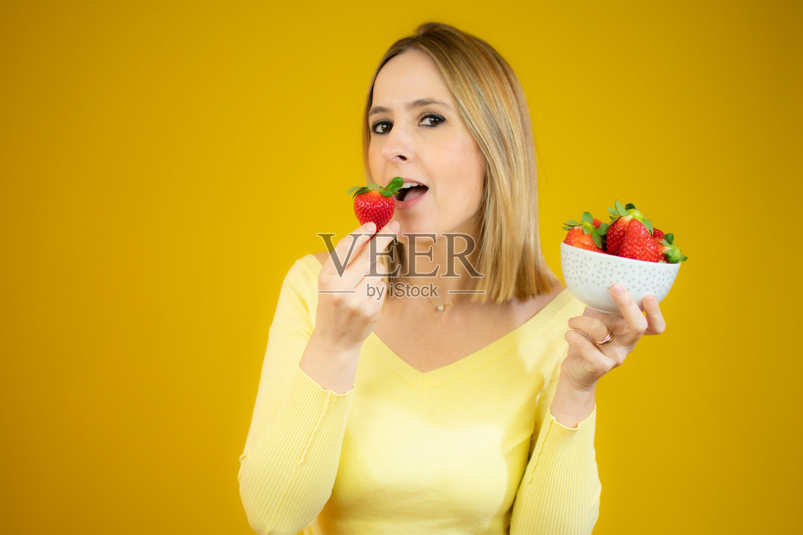 美丽的年轻女子站着，手里拿着一个玻璃碗，黄色的背景上放着草莓照片摄影图片