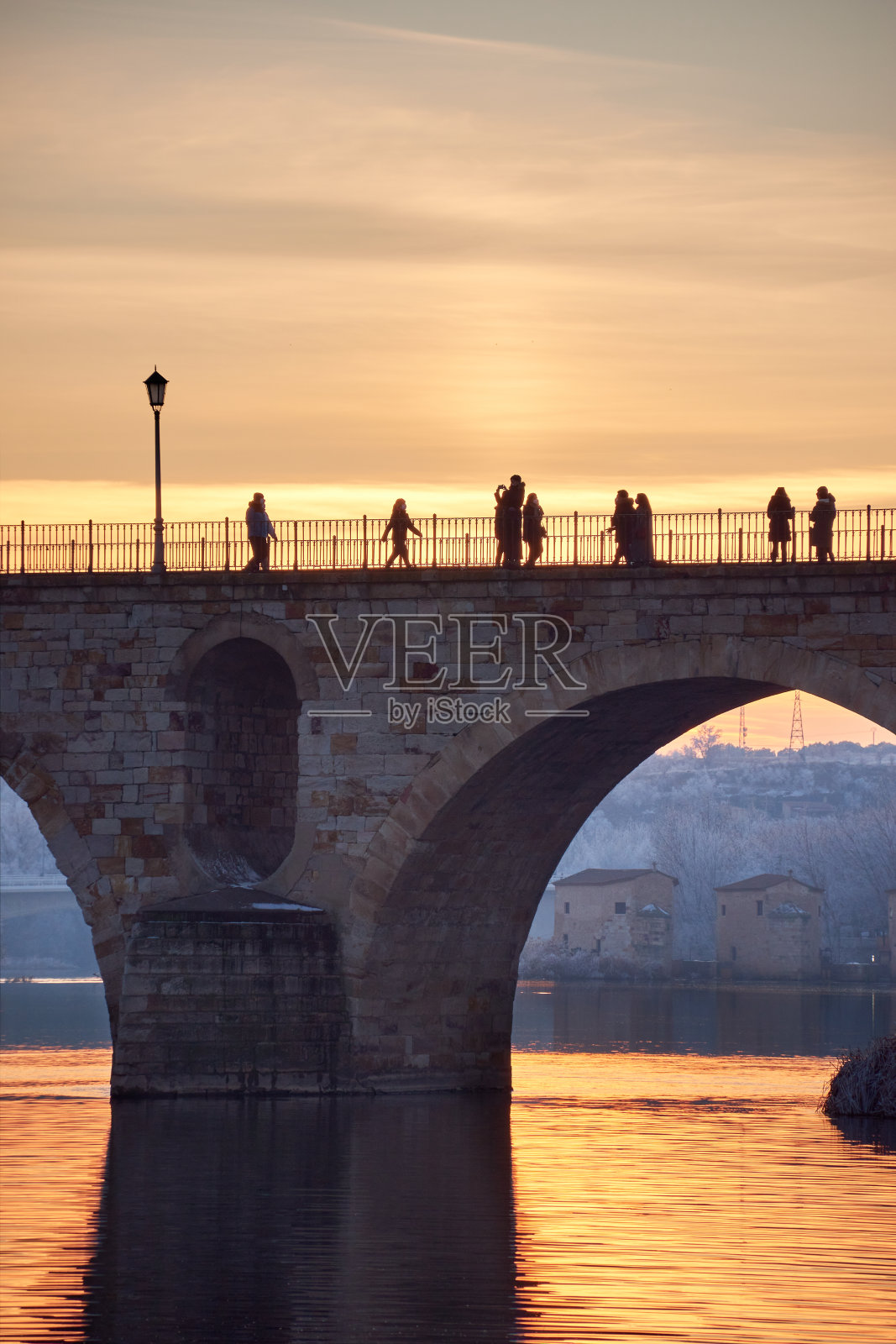 人们在日落时走在石桥上，在冬天的黄金时刻冰冻的树木上。石头拱门，反射着阳光在河上。两座大桥。下水道的背景。俯视图照片摄影图片