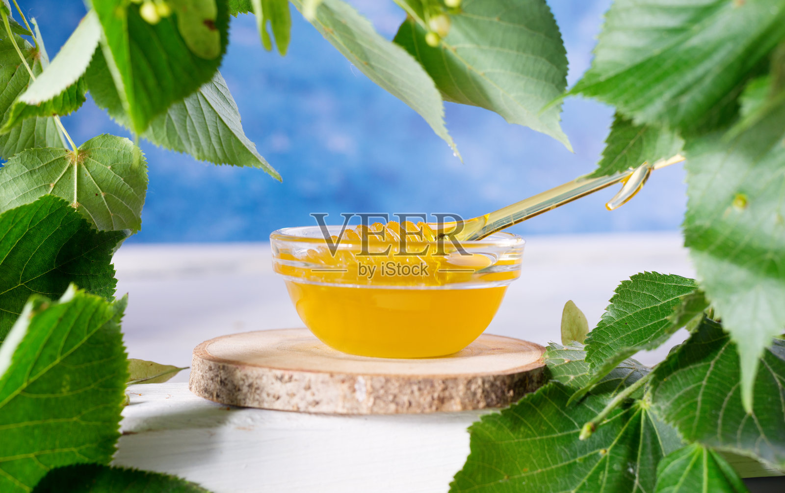 椴树蜂蜜在罐子和碗与蜂蜜勺在一个白色的木制背景。前景中离焦的菩提树枝条。照片摄影图片