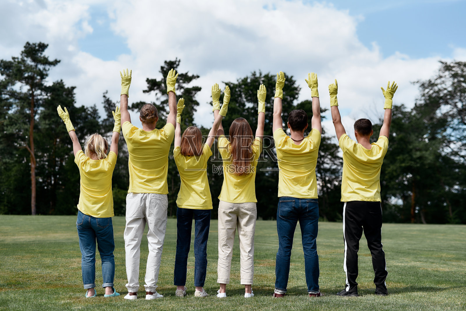 后视图快乐的志愿者穿着黄色制服和橡胶手套举起双手，庆祝成功后清扫森林或公园照片摄影图片