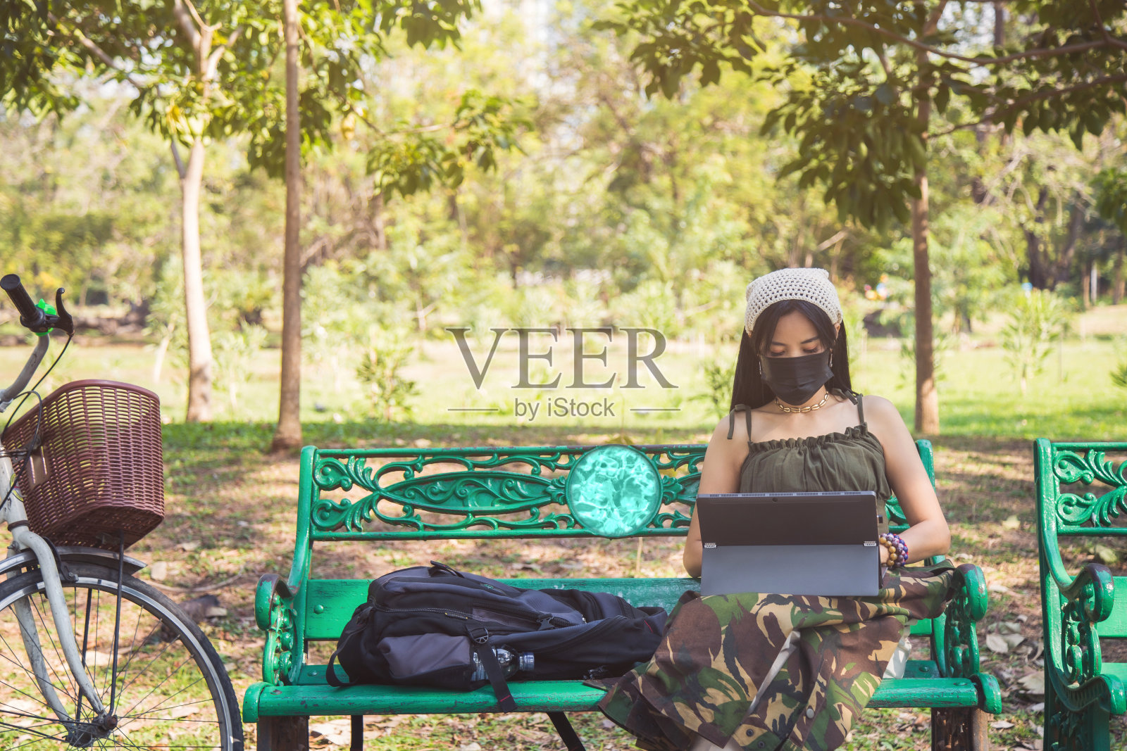 在一个公园里，一名美丽的亚洲女性穿着休闲装，戴着口罩，坐在一辆白色自行车旁的木凳上使用笔记本电脑照片摄影图片