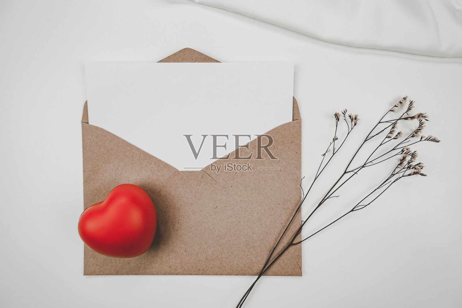 空白白纸放在打开的棕色纸信封上，红色的心和补草干花和白色的布在白色的背景。情人节的概念照片摄影图片