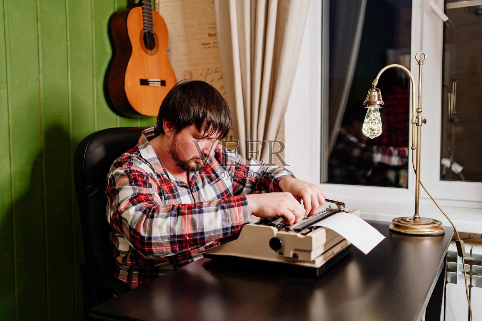 一个穿着格子睡衣的男人晚上在他的家庭办公室里用打字机打字。照片摄影图片