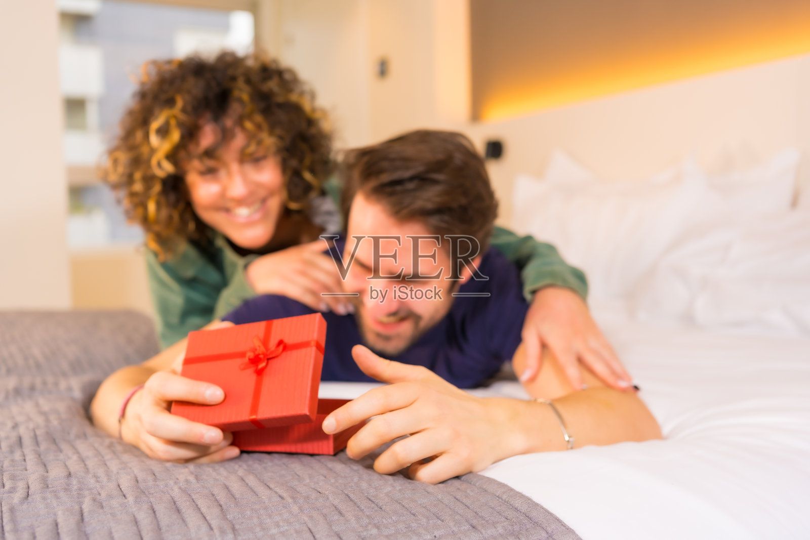 情人节，一对年轻的白种夫妇穿着睡衣躺在床上，新娘用一份漂亮的礼物送给男友照片摄影图片