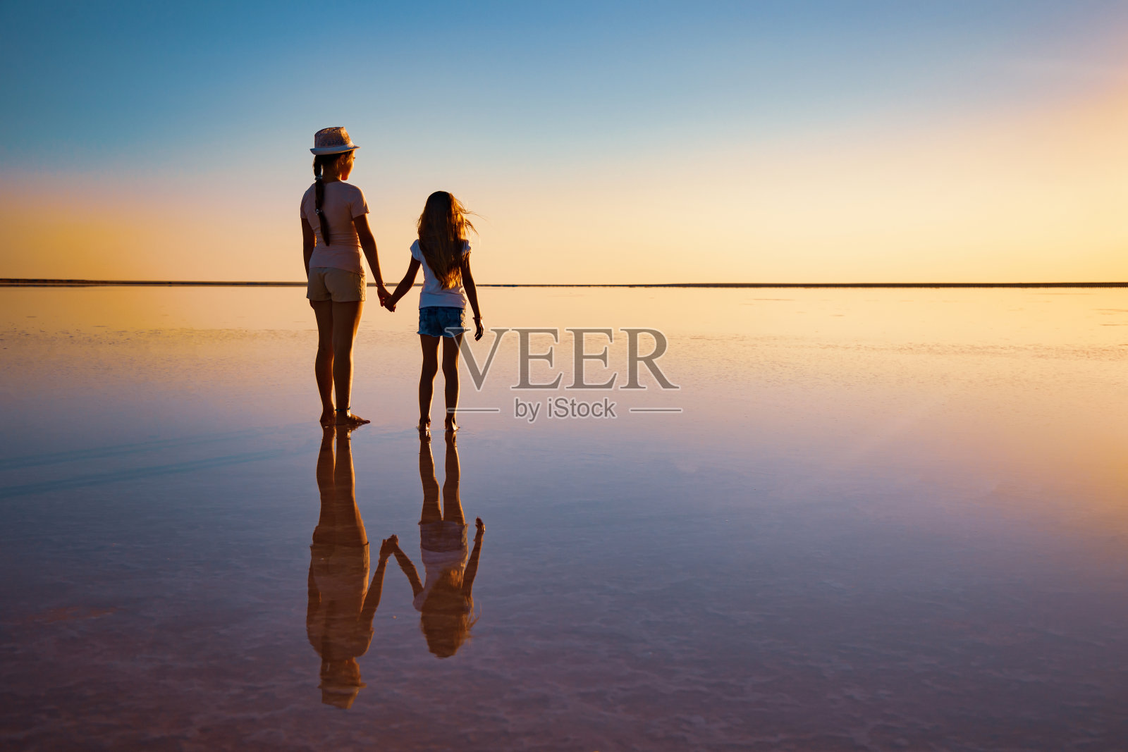 在火红的晚霞中，两个可爱的姐妹沿着镜面的湖散步照片摄影图片