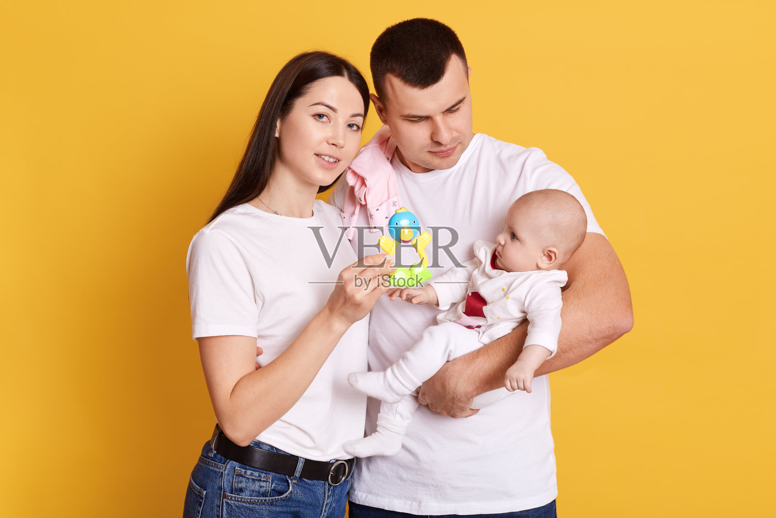 年轻的父母带着孩子，妈妈穿着黑色的长发穿着白色的t恤向孩子展示玩具，爸爸抱着小女儿，一家人孤立地站在黄色的背景下。照片摄影图片