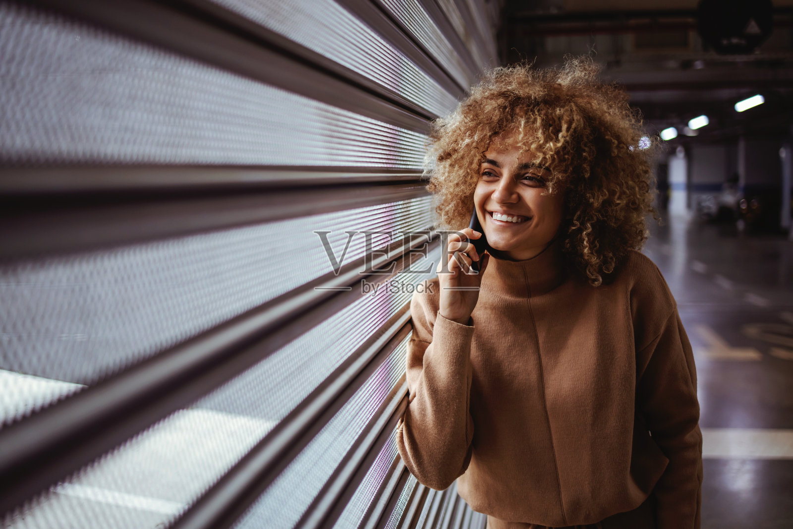 微笑的混血嘻哈女孩靠在车库门上和电话交谈。照片摄影图片