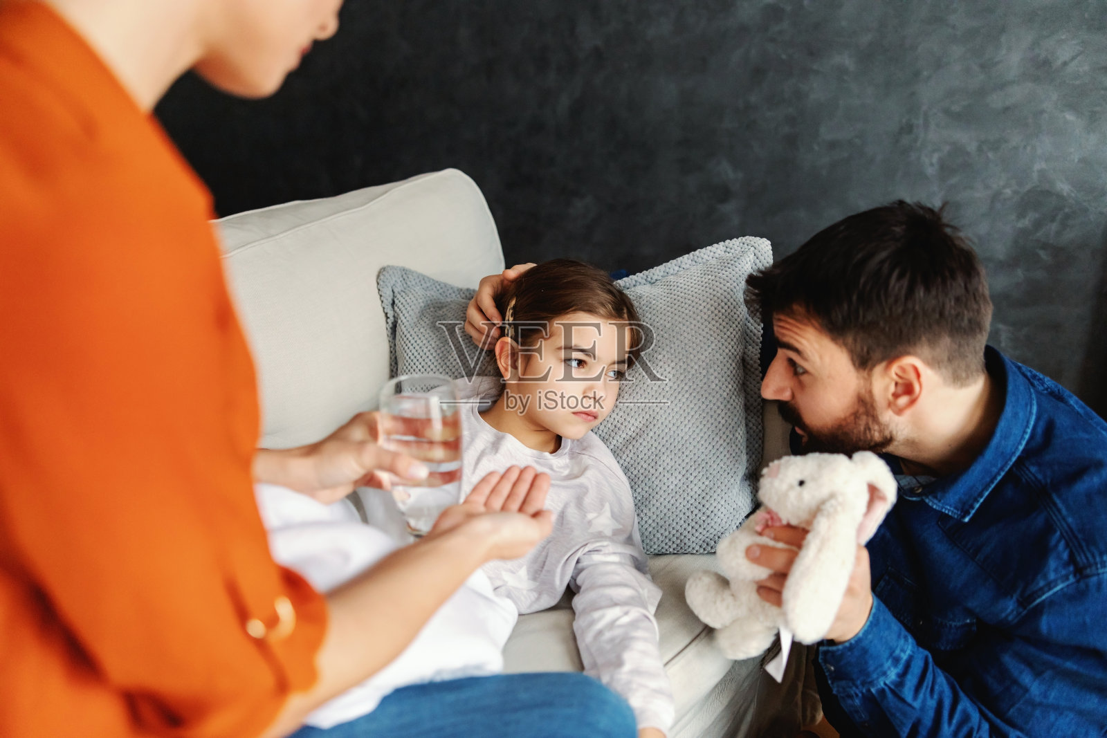 生病的小女孩躺在沙发上，听她父亲说话。母亲正在给女儿吃药和喝水。照片摄影图片