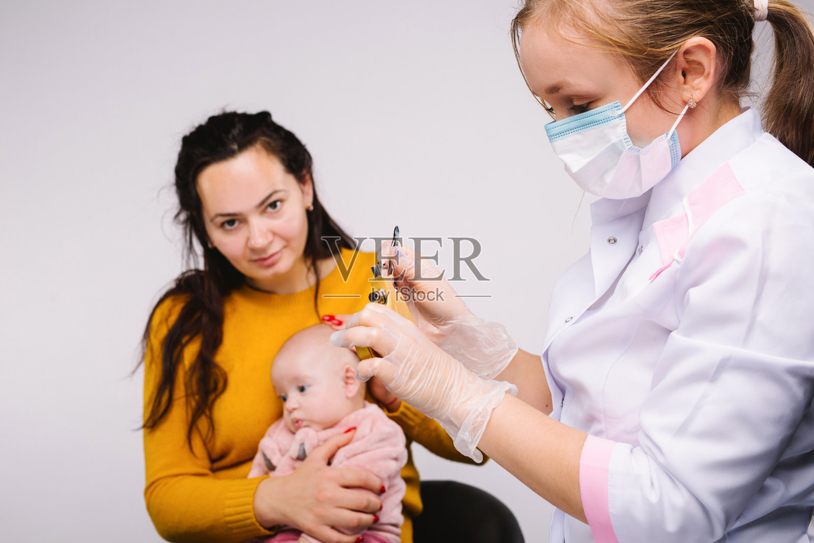 一个医生准备了一把枪和耳环，用来给一个小女孩打耳洞，这个小女孩坐在她母亲的怀抱里。白色背景。照片摄影图片