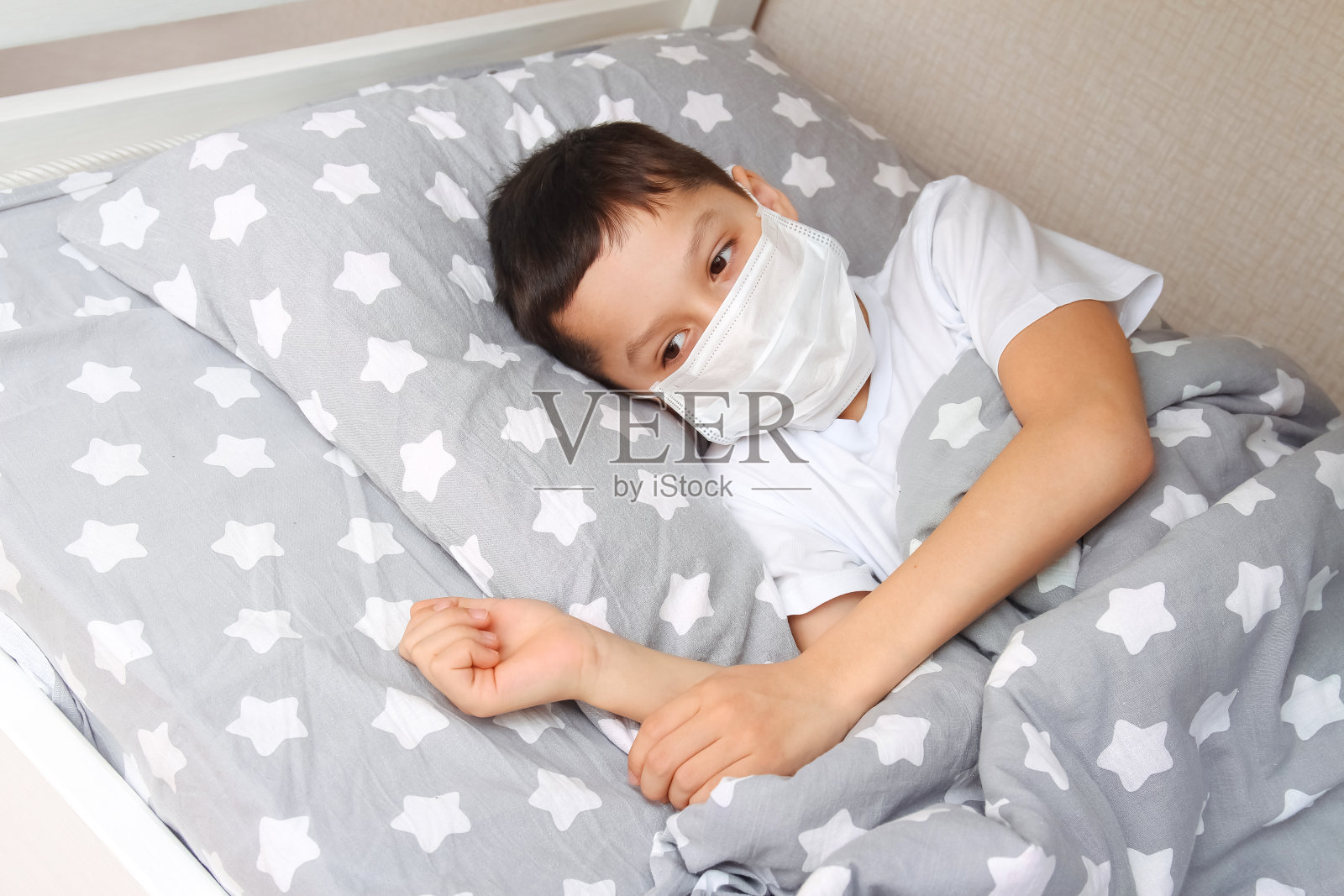 一个黑头发、脸上带着医用口罩的白人男孩躺在房间的床上，盖着一条毯子，他病了。居家治疗的概念照片摄影图片