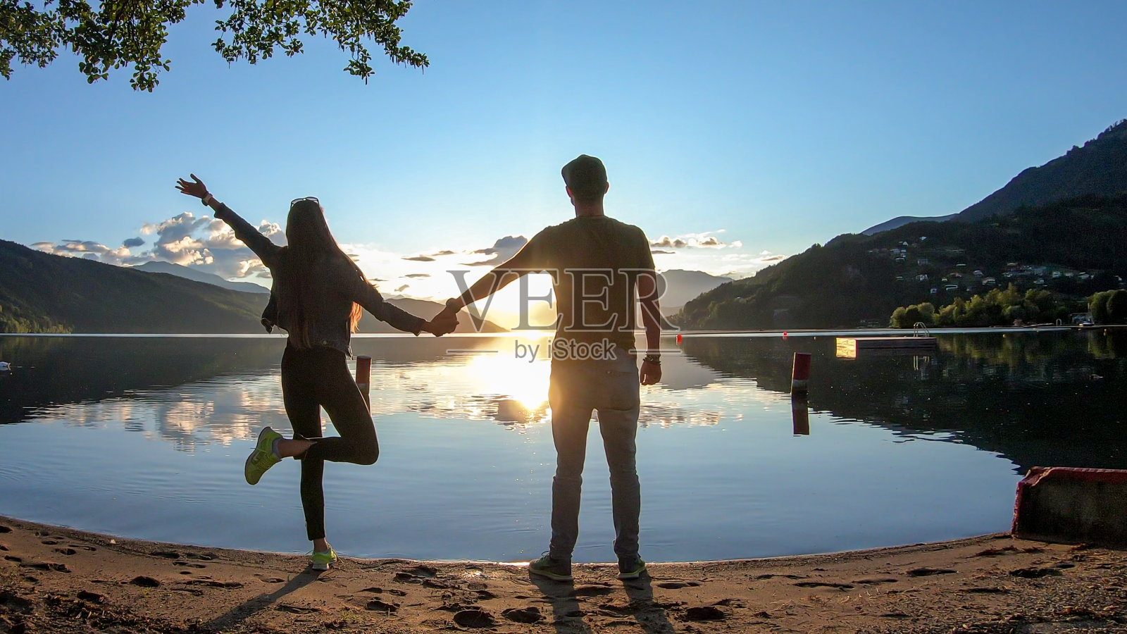 米尔斯塔特湖——一对情侣站在米尔斯塔特湖岸边，在夕阳下手拉手。太阳在高山后面落下照片摄影图片