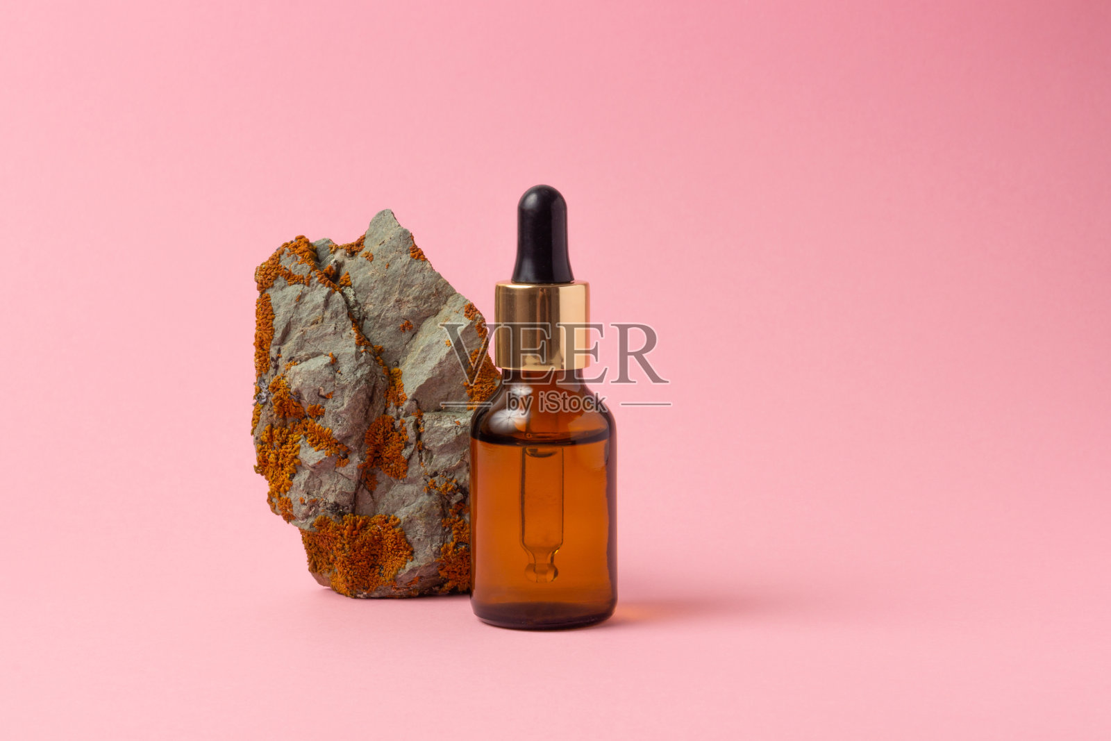 石头旁边放着一个琥珀色的精油和化妆品瓶子。粉红色背景上的玻璃瓶。滴管,喷雾瓶。天然化妆品的概念。照片摄影图片