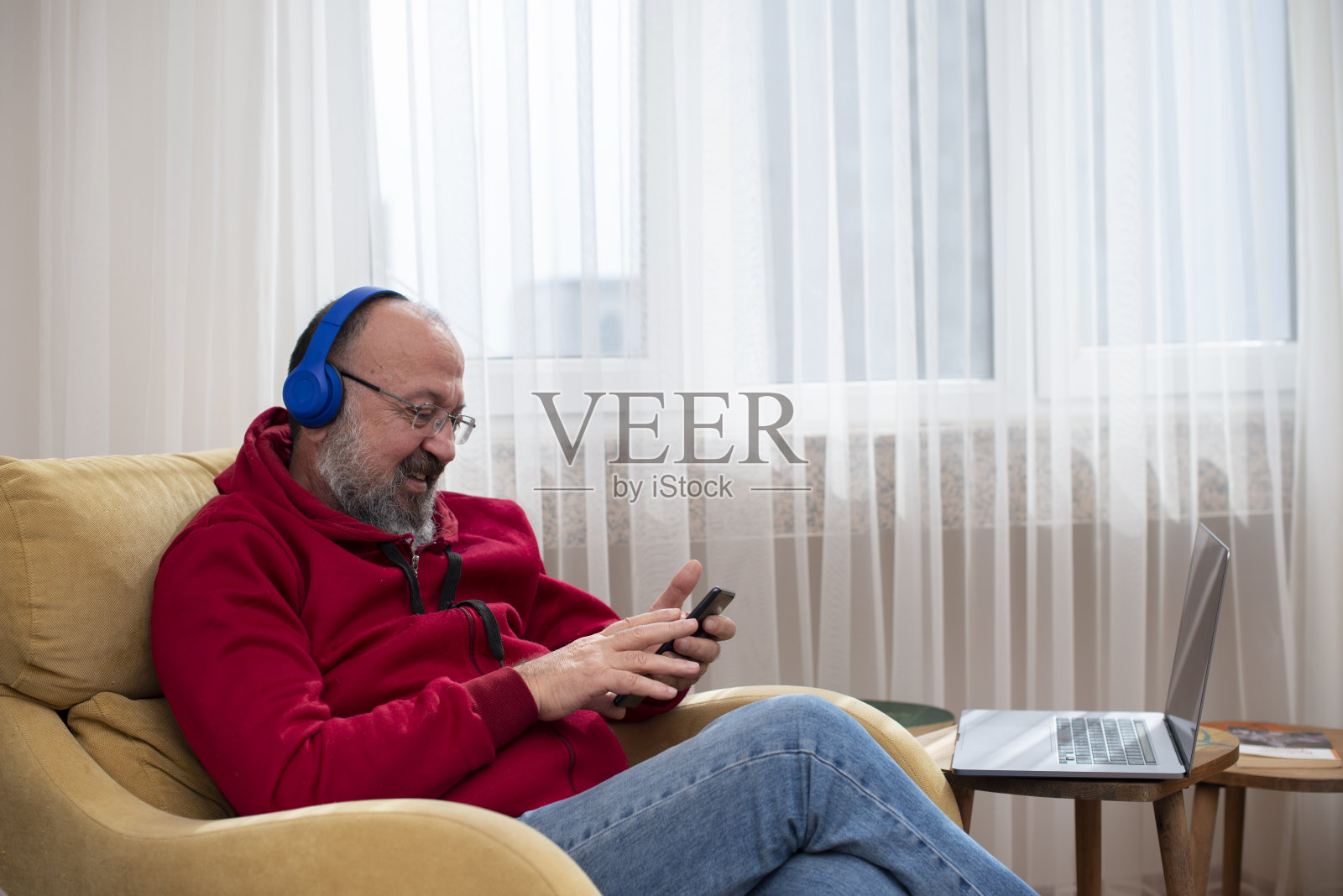 一位退休男子戴着蓝色耳机坐在沙发上听音乐。照片摄影图片