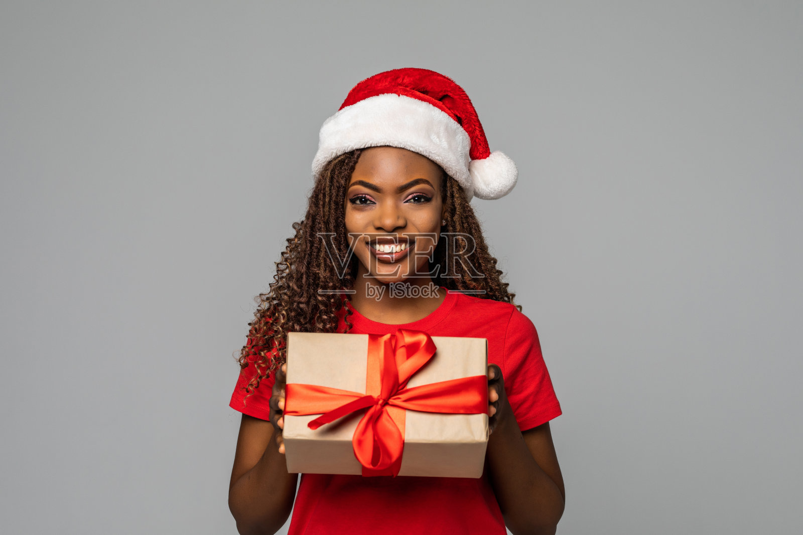 快乐的年轻非洲妇女在圣诞老人的帽子拿着圣诞礼物，而站在灰色的背景照片摄影图片