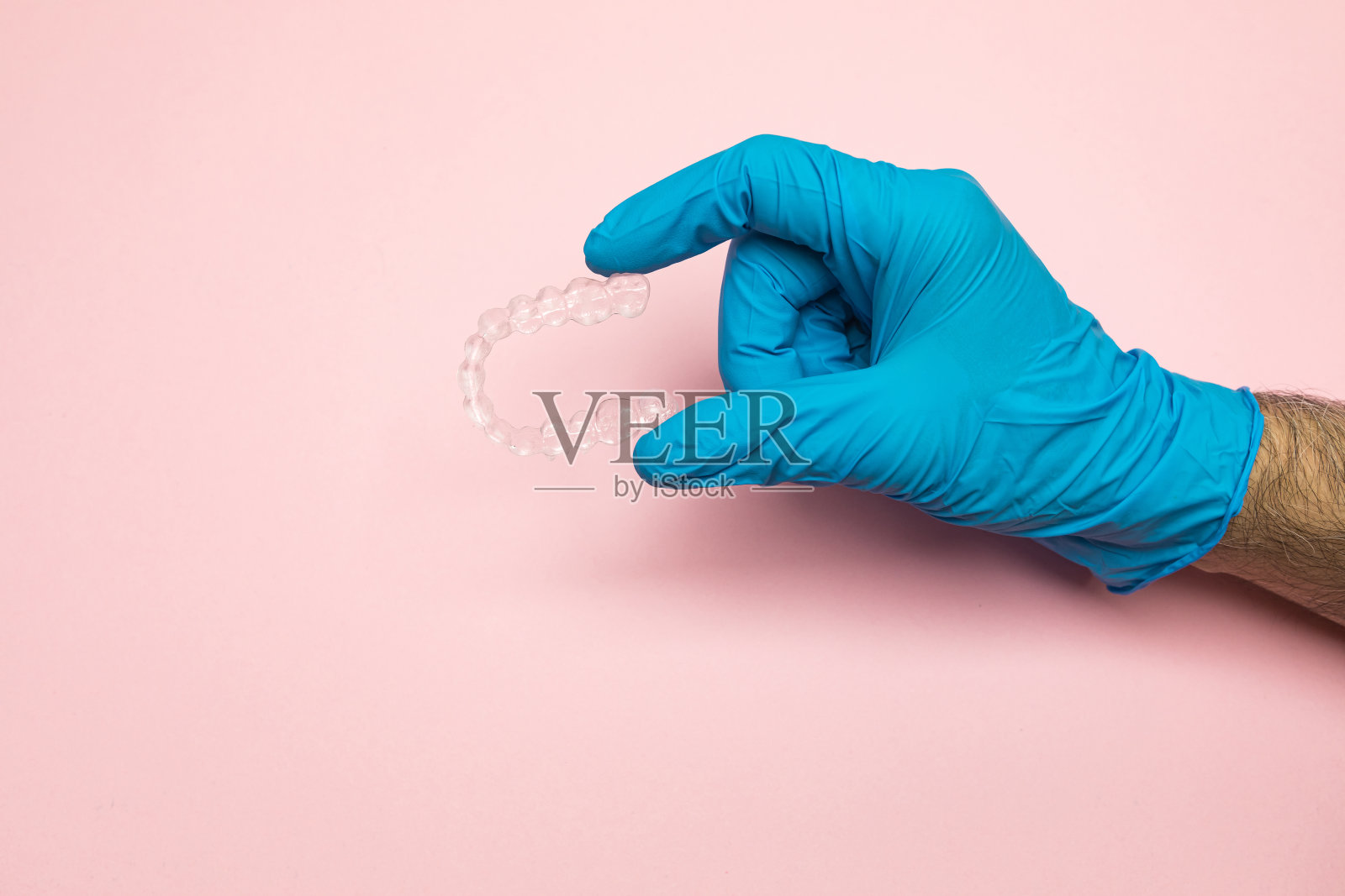 牙医戴着蓝色乳胶手套的手在粉红色的背景上持有一个清晰的牙齿对准器。牙套牙概念照片摄影图片