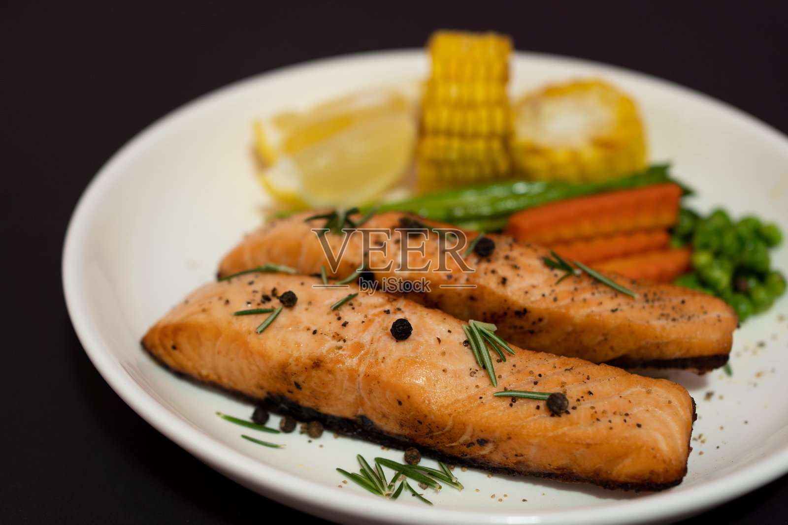 两片鲑鱼排配蔬菜，烤玉米，芦笋，胡萝卜，青豆，柠檬，胡椒在黑色的背景上。照片摄影图片