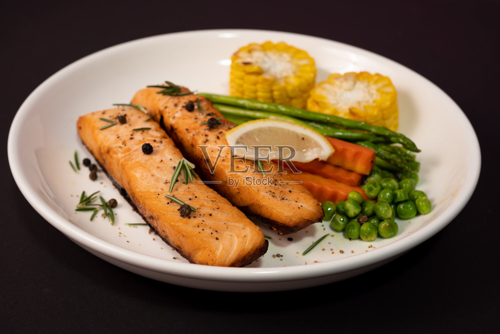 两片鲑鱼排配蔬菜，烤玉米，芦笋，胡萝卜，青豆，柠檬，胡椒在黑色的背景上。照片摄影图片