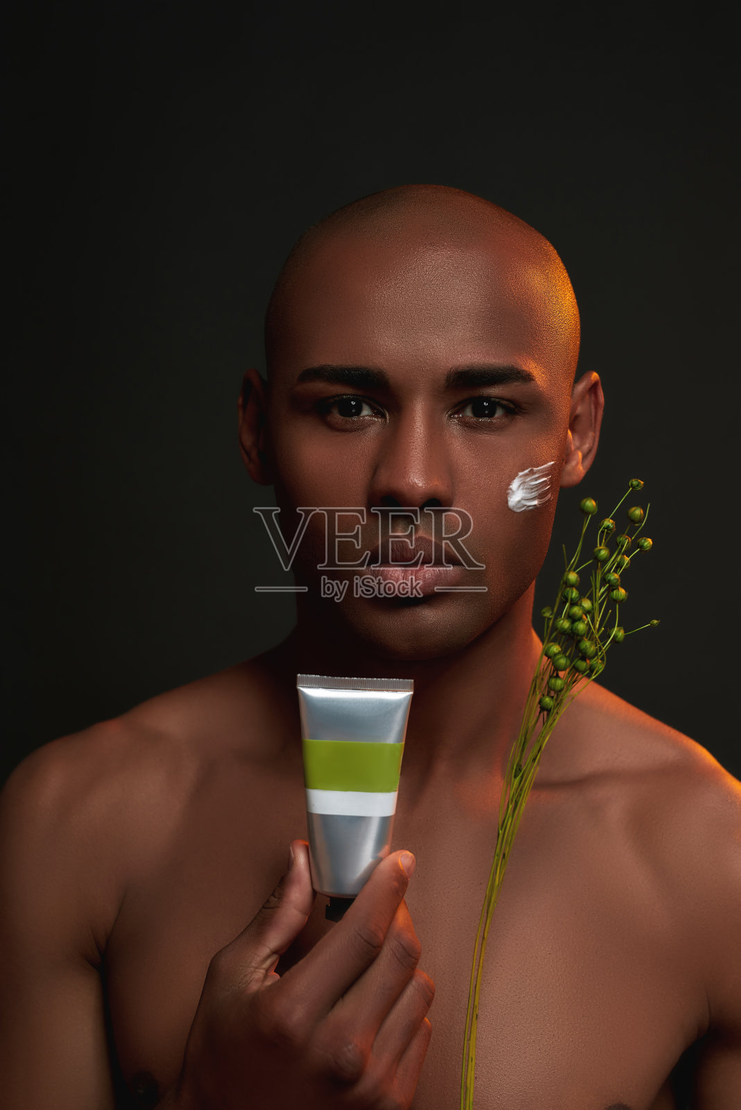 赤裸上身的非洲裔美国人的肖像看着相机，拿着自然的有机美容产品，与霜应用在他的脸颊孤立的黑色背景照片摄影图片