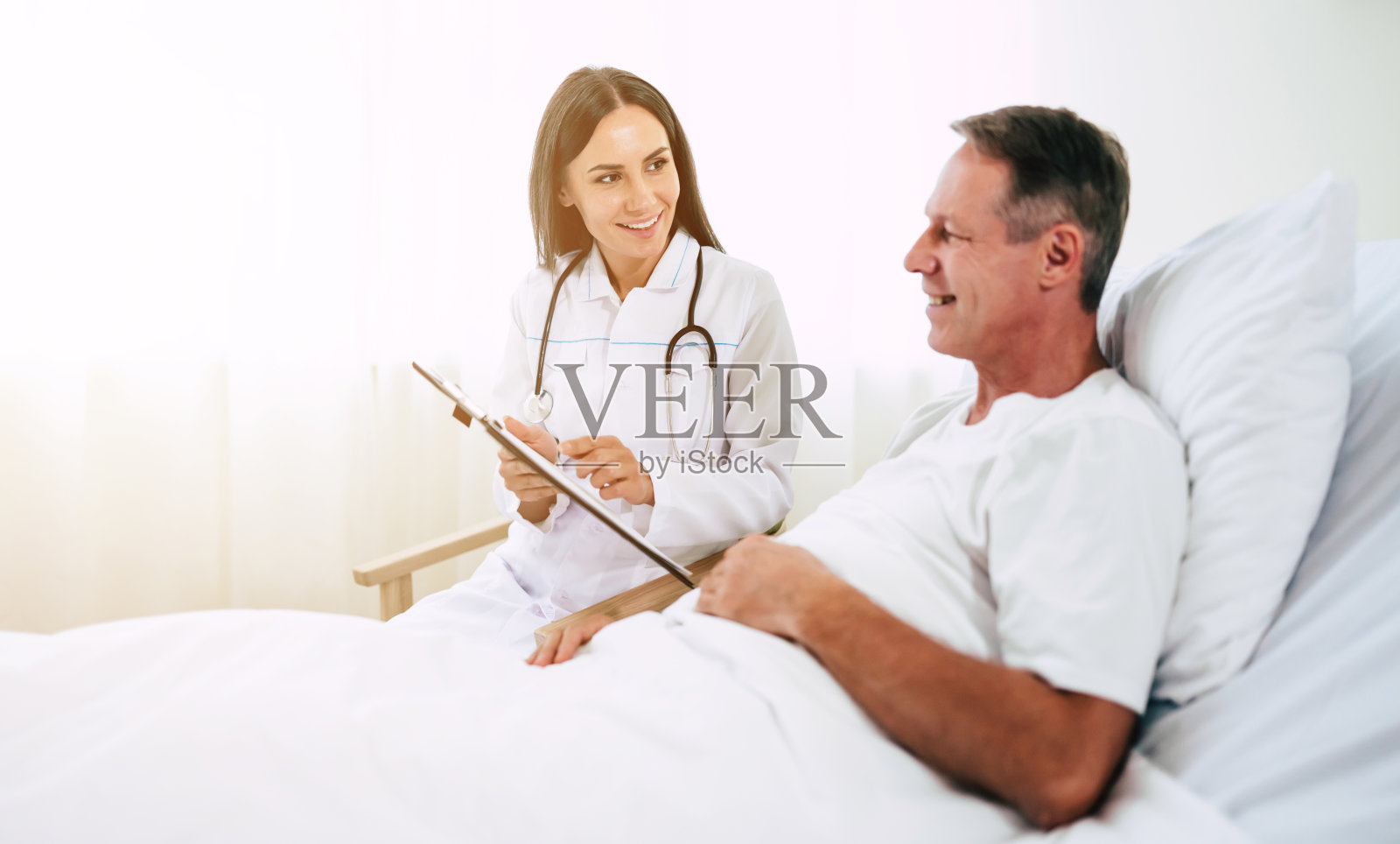 英俊成熟的病人正躺在病床上，与一位自信的年轻白大褂女医生交谈照片摄影图片