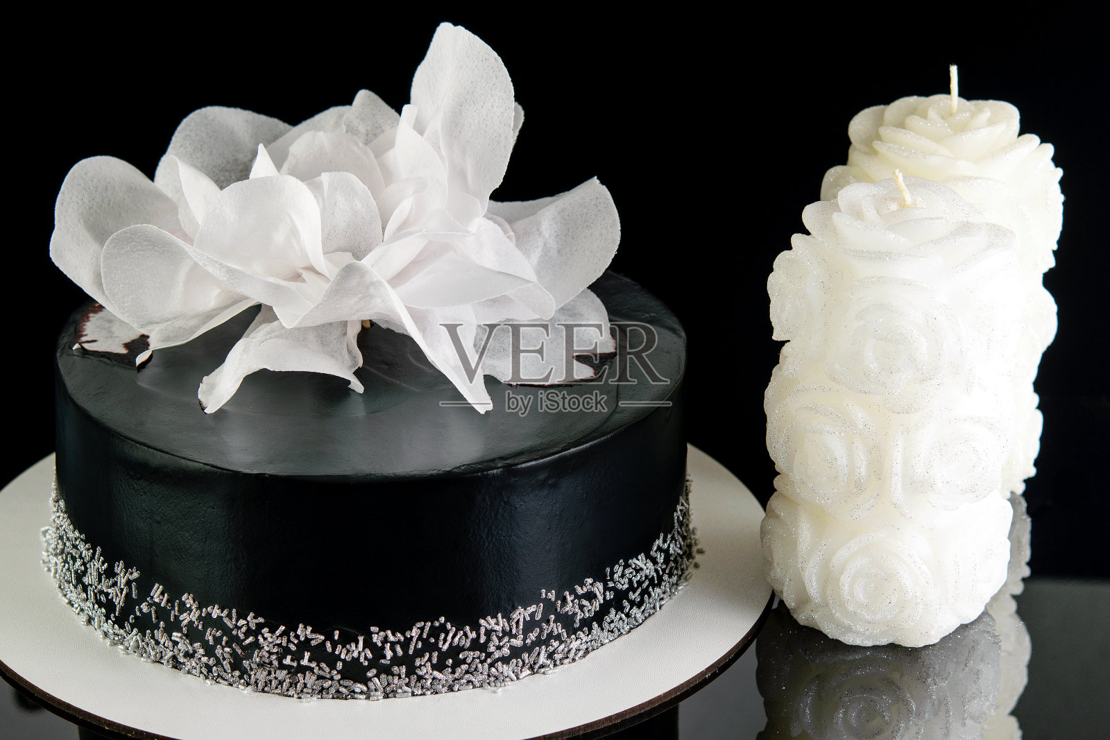 美味的甜点。自制的节日黑色蛋糕，装饰着白色的大花，在黑色的反光背景上插着白色的蜡烛。照片摄影图片