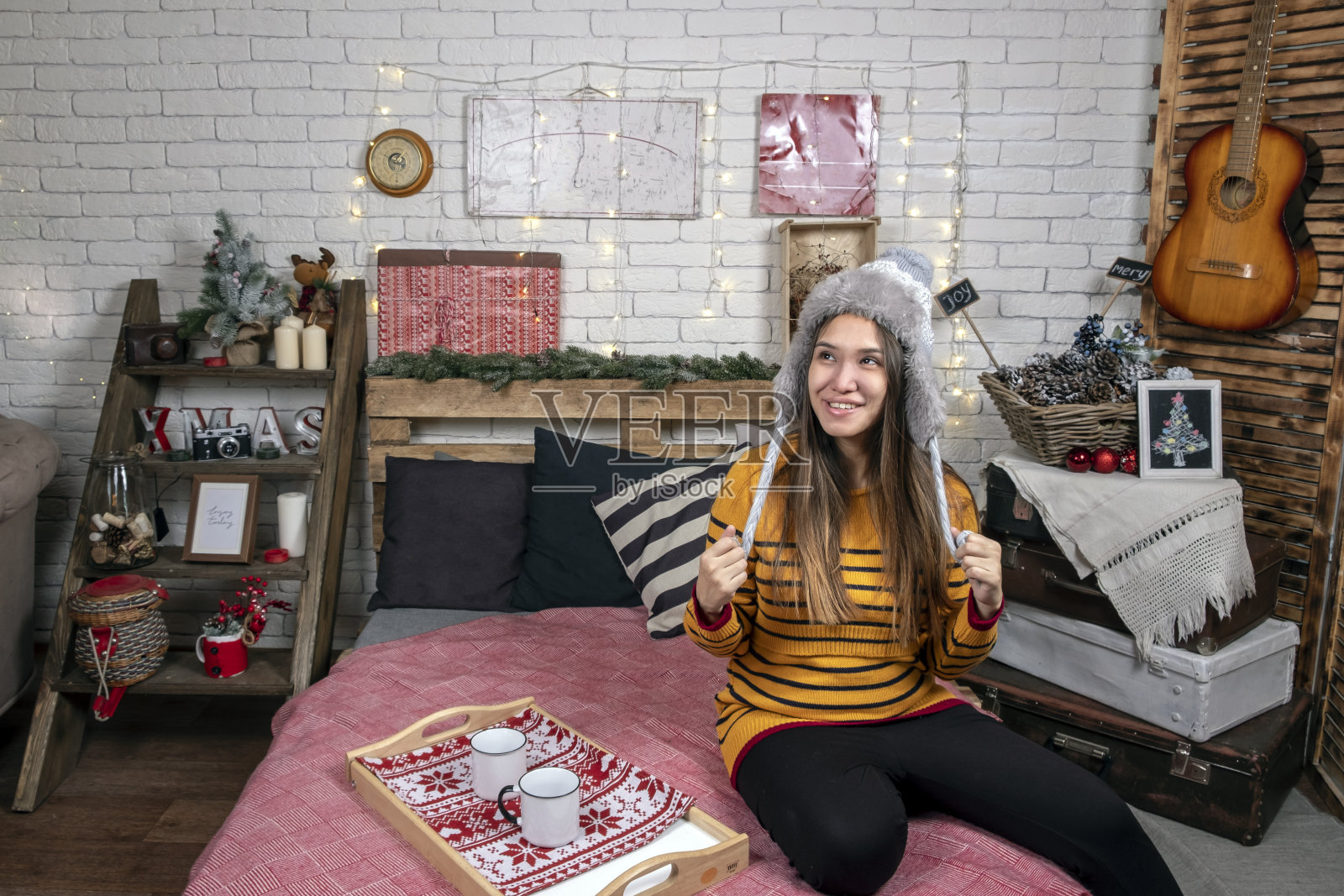 快乐的年轻亚洲妇女在帽子是坐在床上杯咖啡或茶在舒适的圣诞节装饰卧室附近。圣诞假期。照片摄影图片