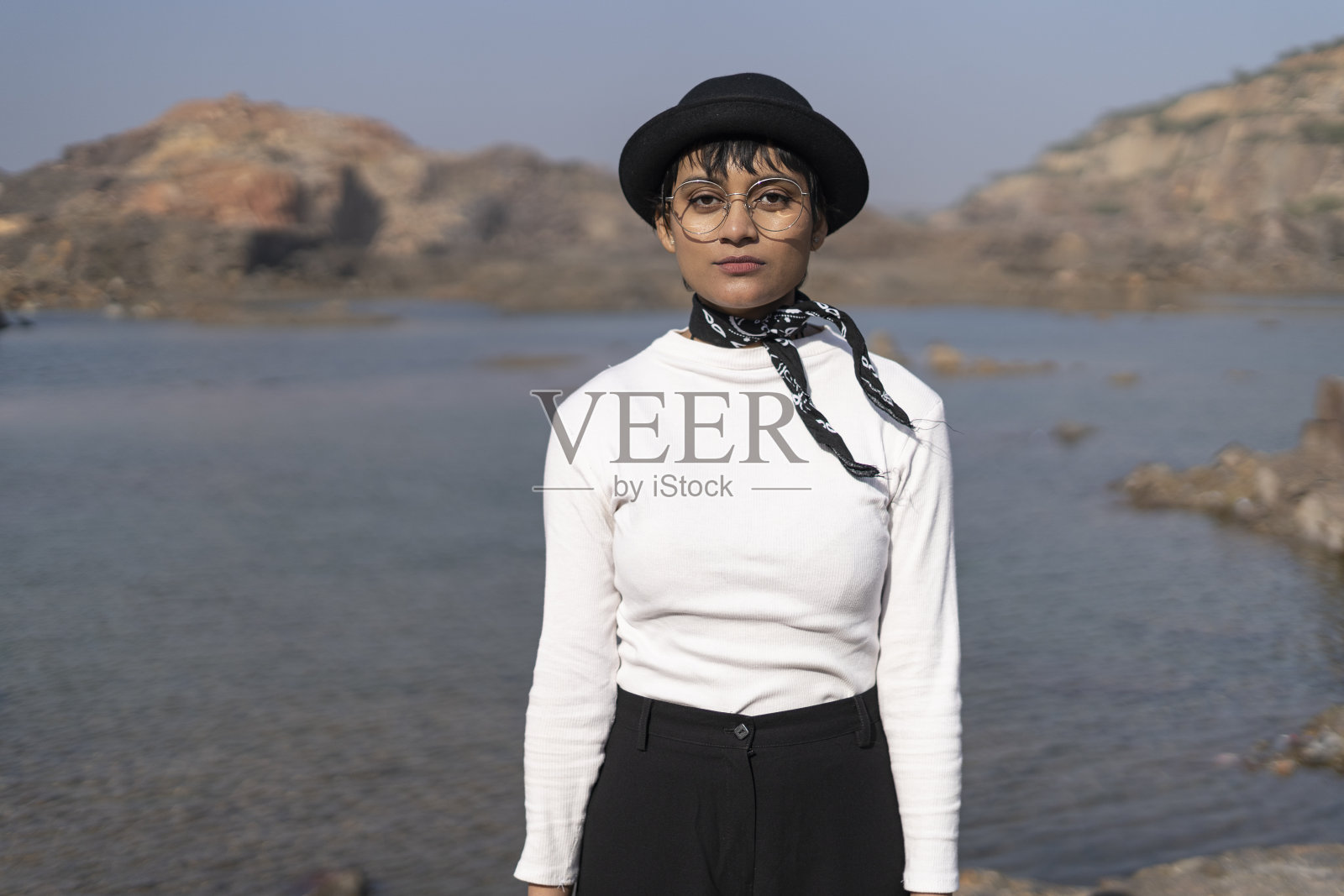 一个印度女孩穿着白色上衣和黑色裤子，站在群山环绕的湖边。照片摄影图片