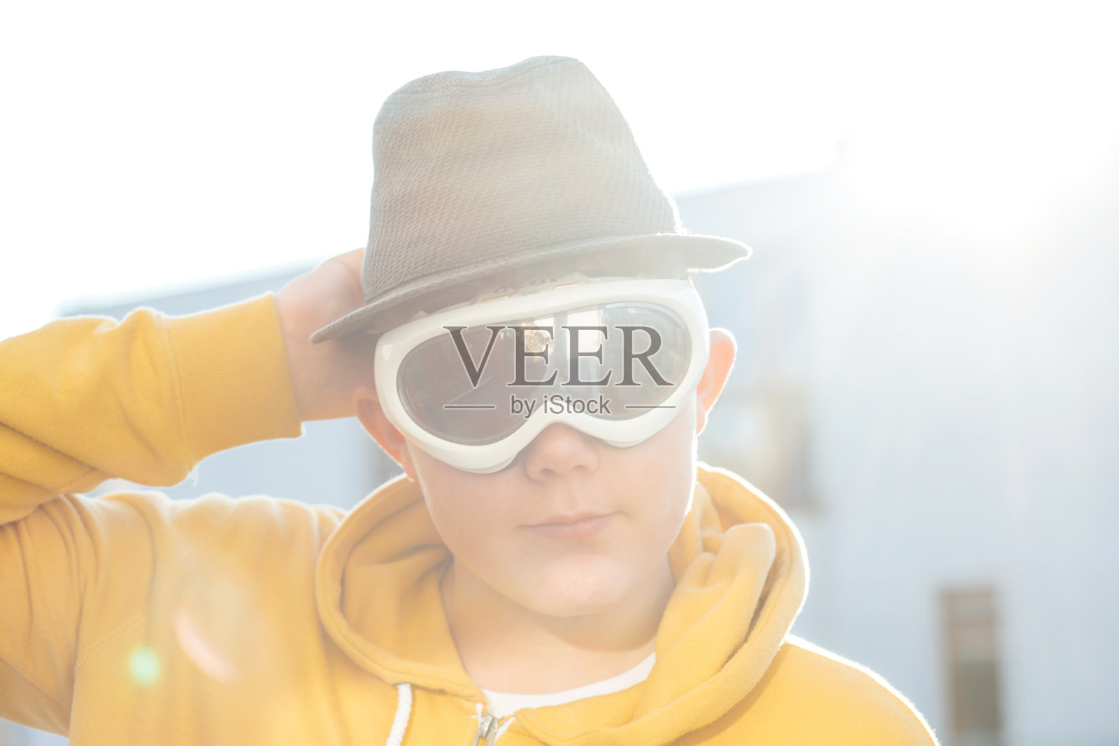 滑稽的肖像，大头照——一个十几岁的男孩手握软呢帽，戴着运动护目镜，穿着黄色连帽衫，站在居民区外背光下，严肃地扮演角色。照片摄影图片