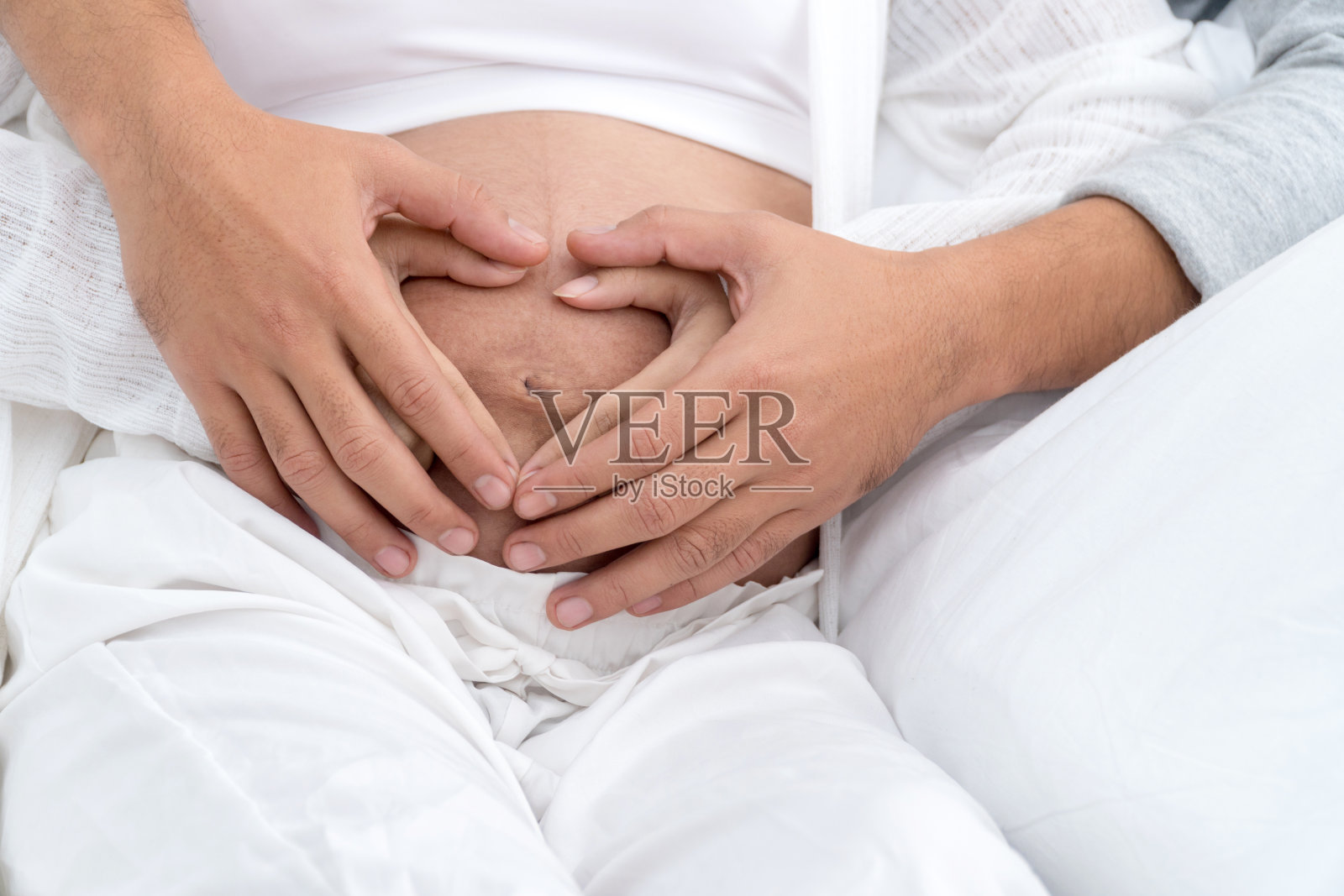 一对幸福的夫妇用手在怀孕的肚子上画心形。一名孕妇和她的丈夫将他们的手做成心形放在她怀孕的婴儿上。期待孩子，爱，关心的概念。照片摄影图片