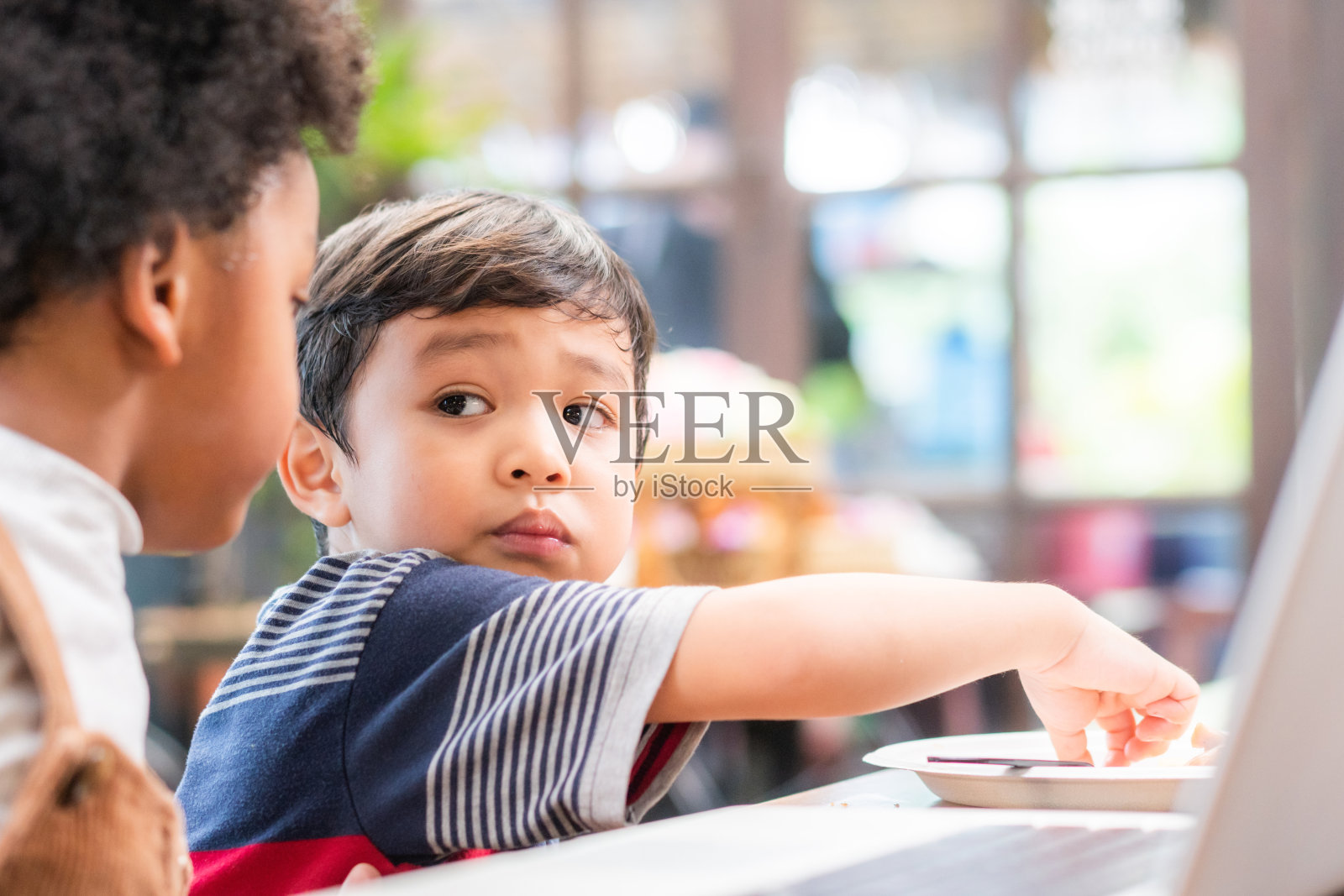 多样性小孩一起在电脑上看电影。亚洲男孩捡起小吃，并与黑人男孩在餐厅或咖啡馆交谈照片摄影图片