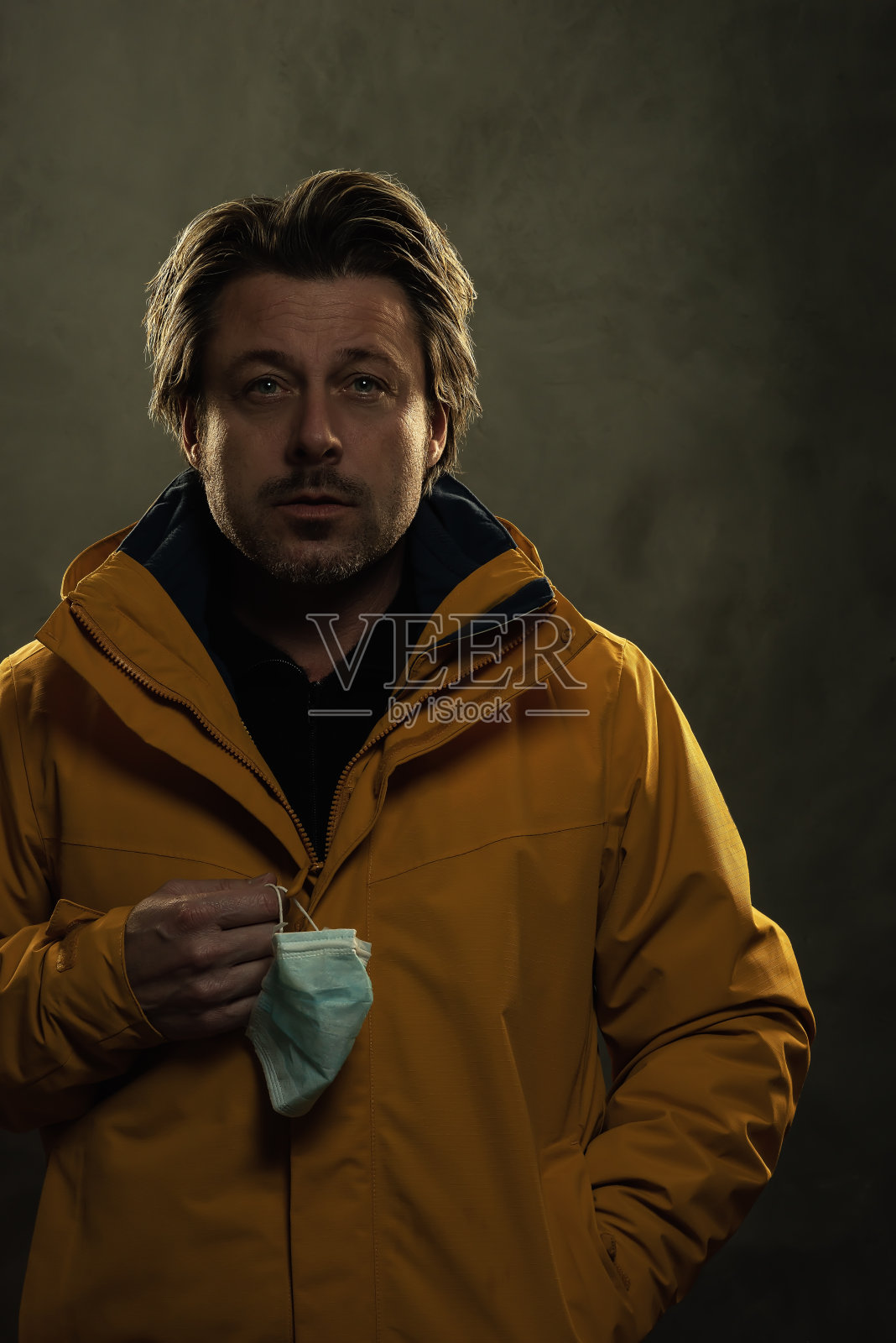 灰色墙壁前，一个金发男子拿着卫生口罩，穿着一件黄色外套。照片摄影图片