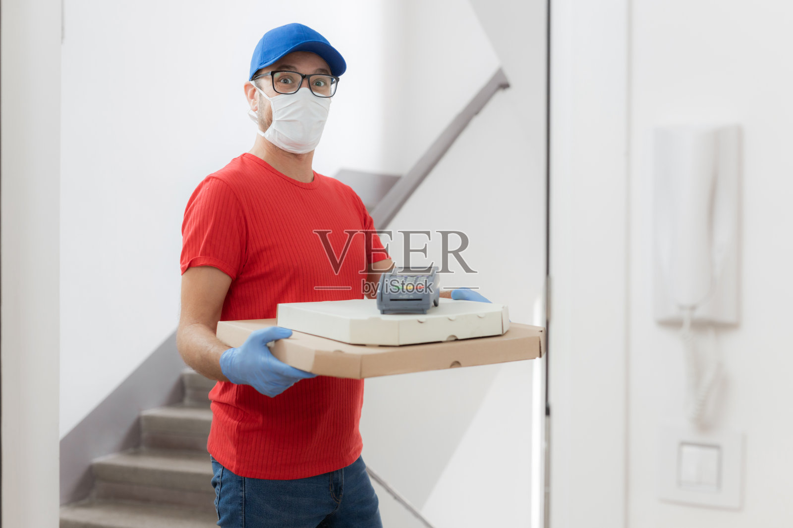 送餐员戴着防护面具，拿着披萨盒和非接触式POS机站在门口。照片摄影图片