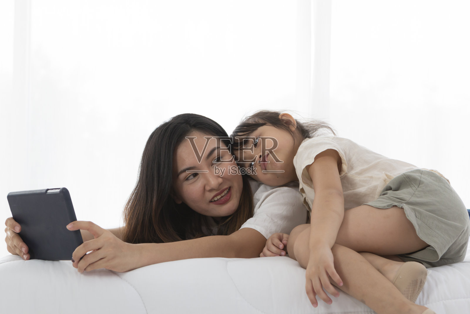 小女孩和妈妈在床上玩，她躺在妈妈背上看着平板电脑屏幕。年轻的母亲和女儿在床上一边玩平板电脑一边聊天。照片摄影图片