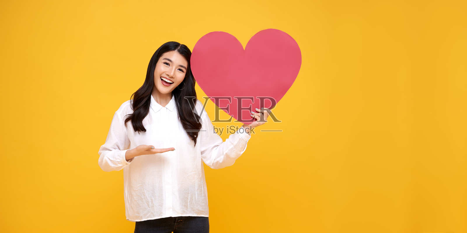 年轻美丽的亚洲女人显示红心标志孤立在黄色背景。爱和快乐的情人节概念。照片摄影图片