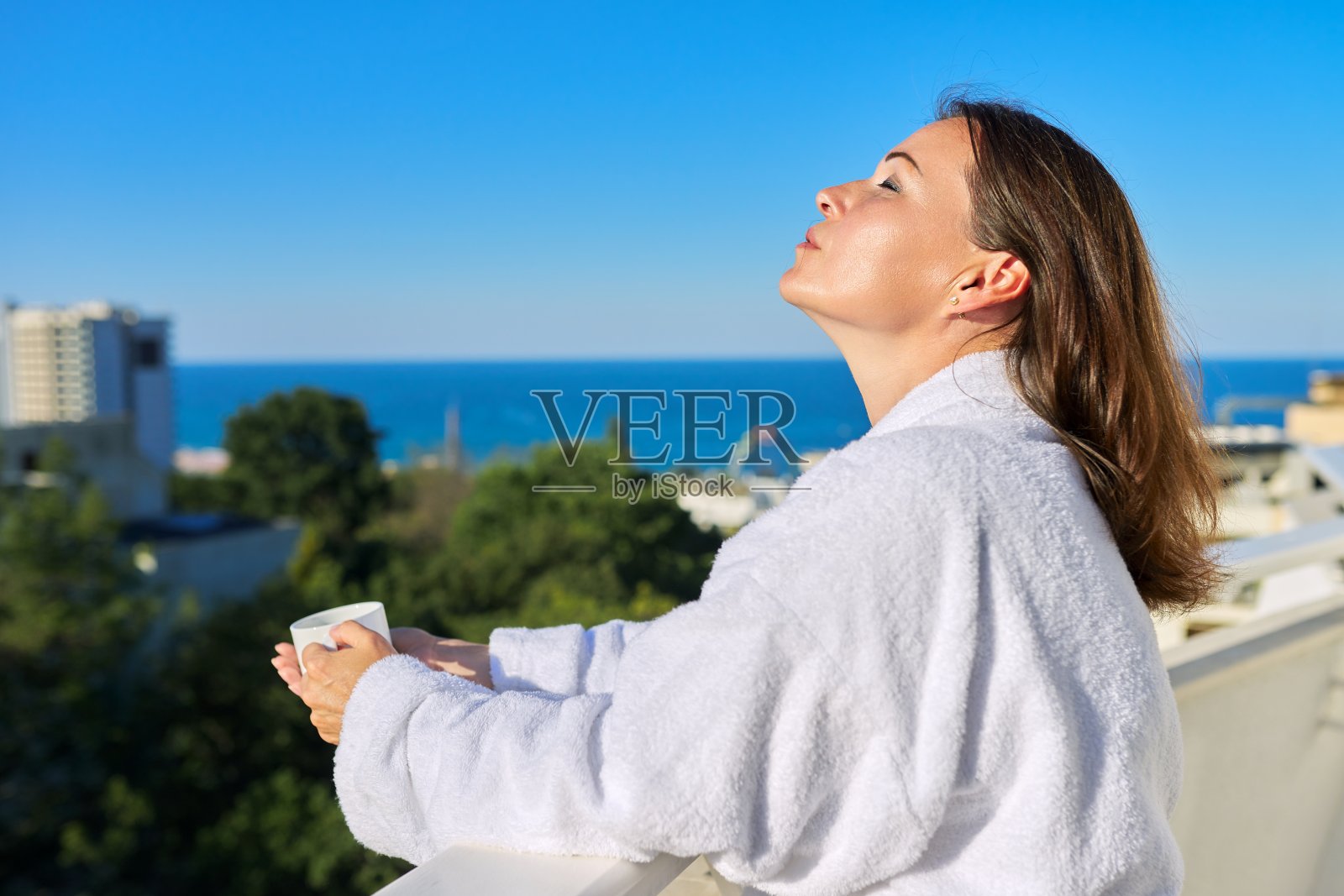 穿着白色浴衣的快乐成熟女性在海滨度假小镇酒店的阳台上照片摄影图片