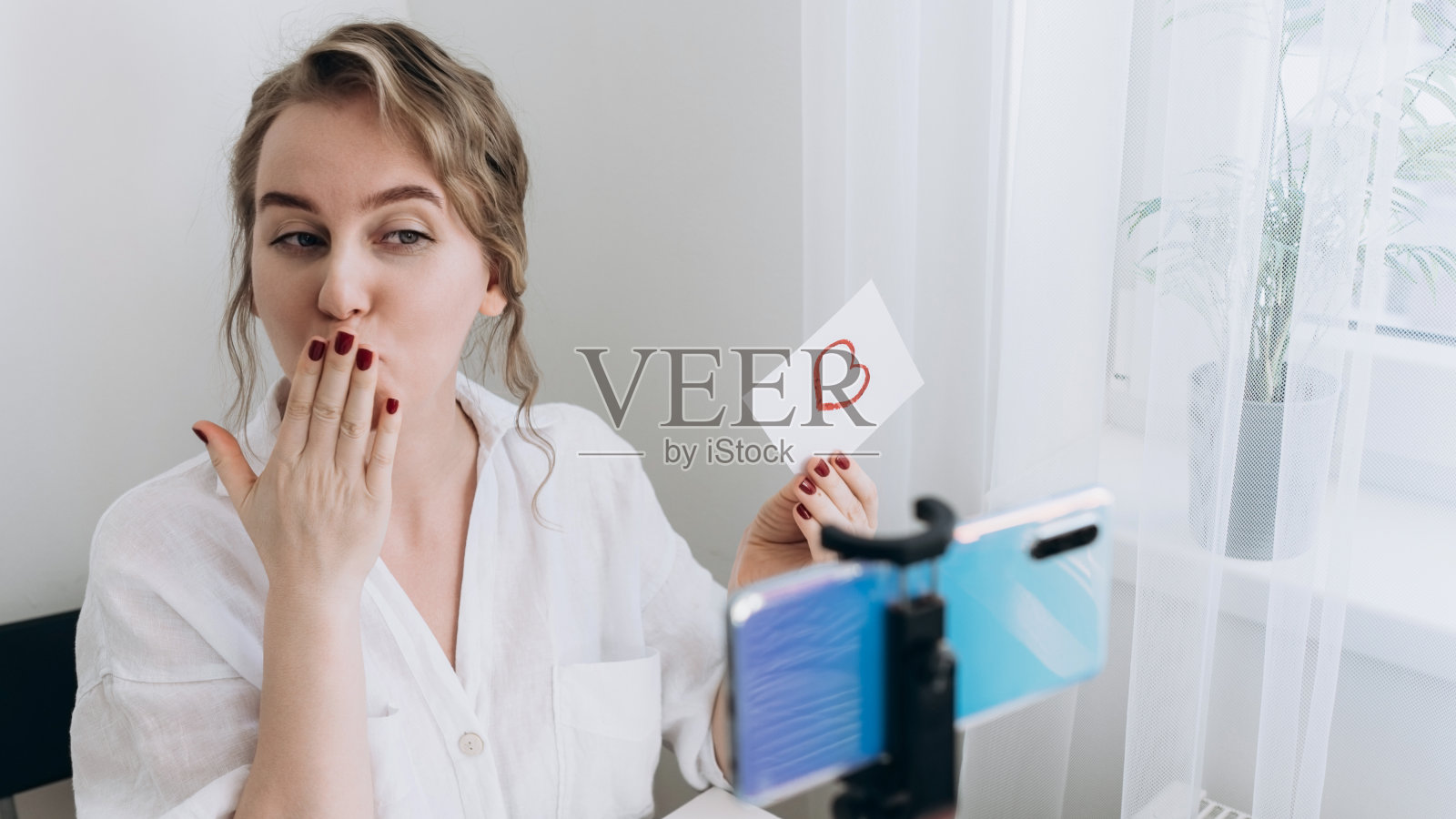 一名女子在用智能手机进行视频通话时，展示了一幅心形的图画，并飞吻。遥远的相识还是隔离期间的爱情宣言。虚拟情人节照片摄影图片