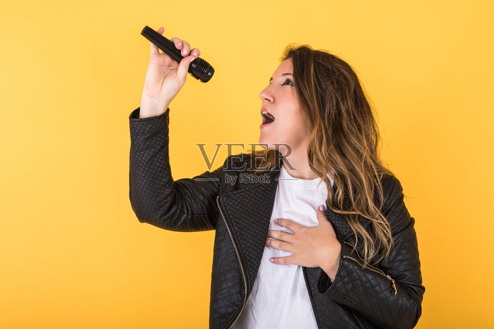 年轻的歌手女孩穿着黑色皮夹克唱歌与麦克风在黄色的背景。照片摄影图片