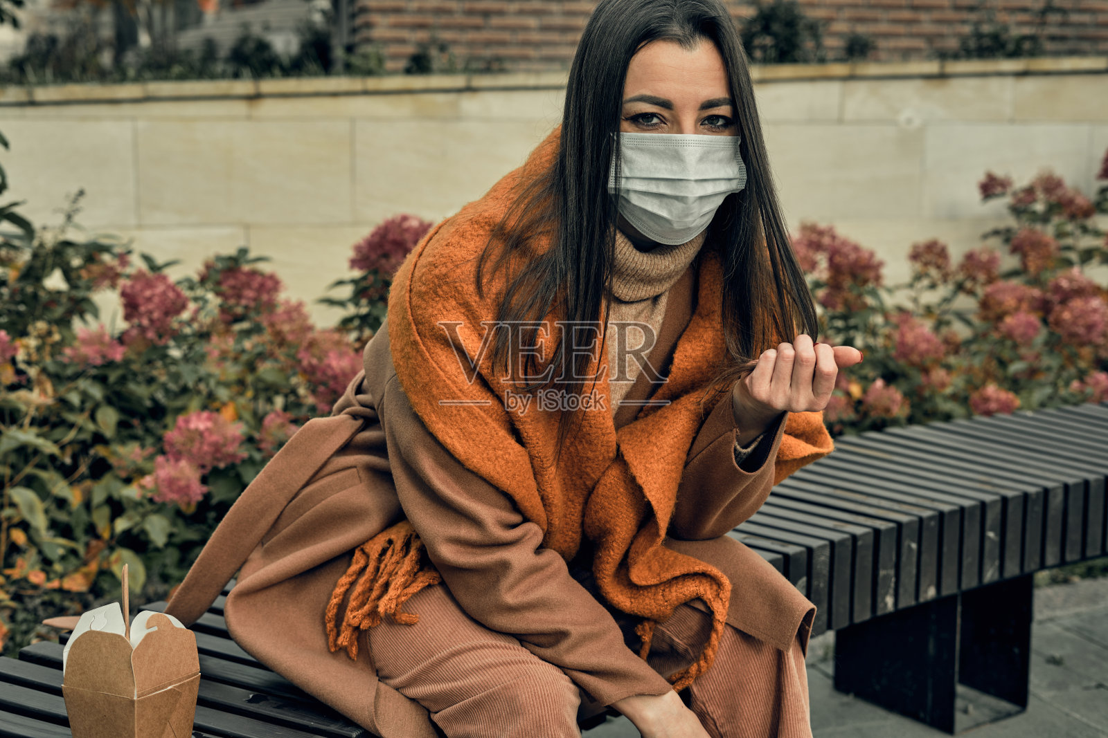 一个戴着防护面具的女孩坐在公园的长椅上，手里拿着一盒炒面。使用环保包装。城市里的快餐流行照片摄影图片