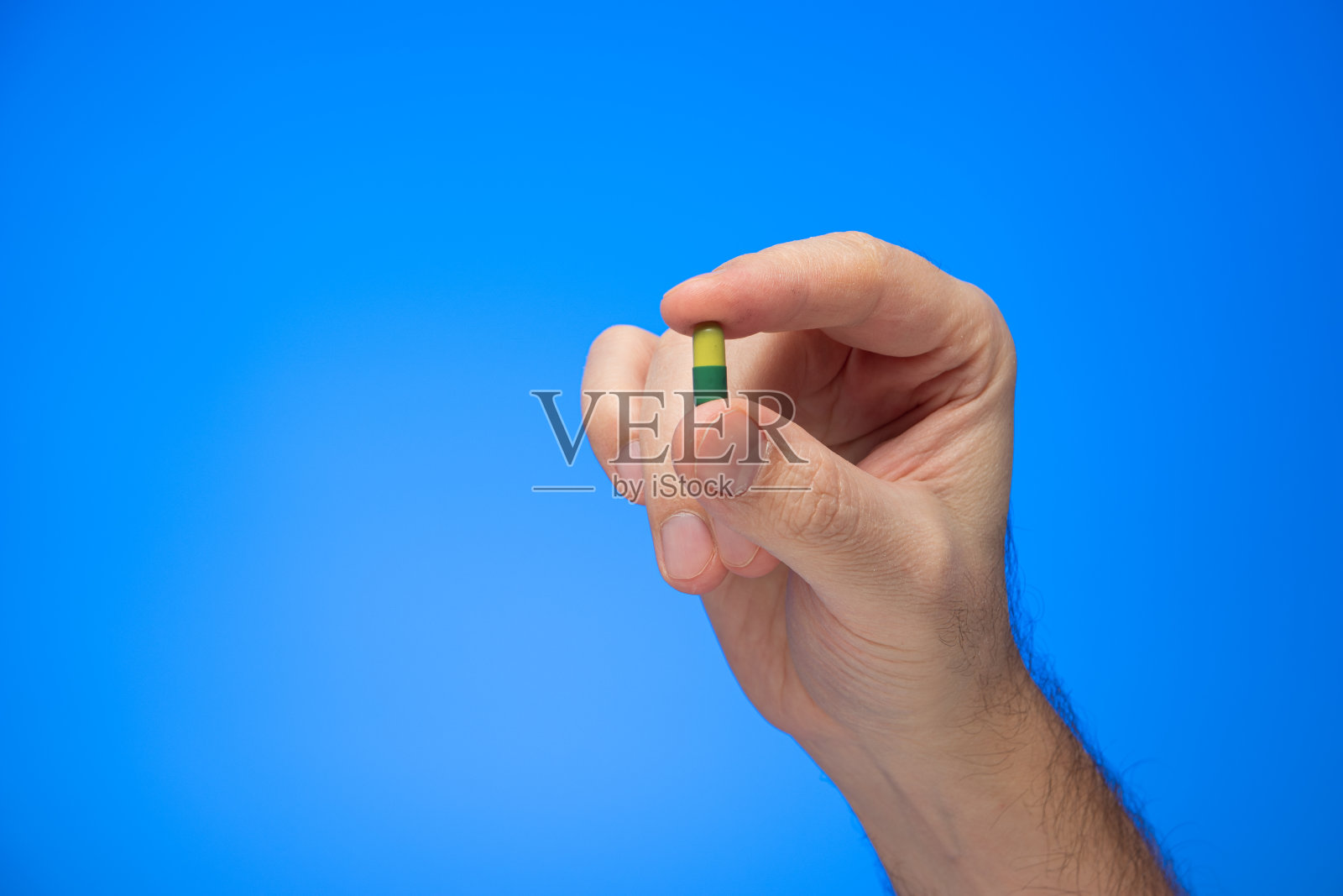 黄色和绿色的胶囊药夹在手指之间，由白人男性的手孤立在蓝色的背景上拍摄照片摄影图片