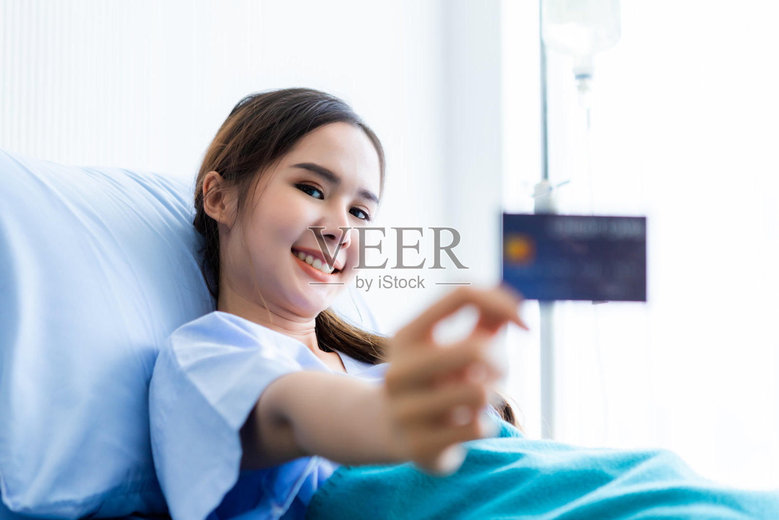 亚洲年轻女病人笑脸上展示着拿着信用卡躺在病房床上医院的背景，支付医疗费的概念照片摄影图片