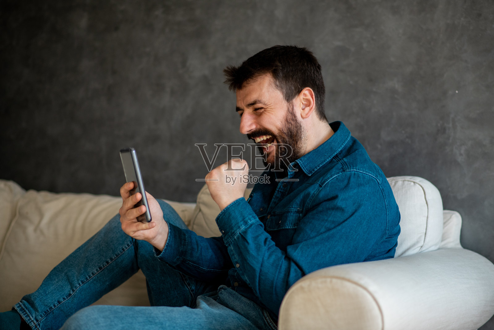 大胡子年轻人边笑边读短信。他坐在客厅的沙发上。照片摄影图片