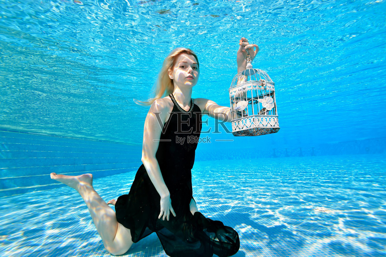 一个金发女孩在水下的时尚肖像。她穿着黑色裙子，手拿白色格子裙，在室外游泳池里摆姿势。明亮的颜色。水下摄影。照片摄影图片