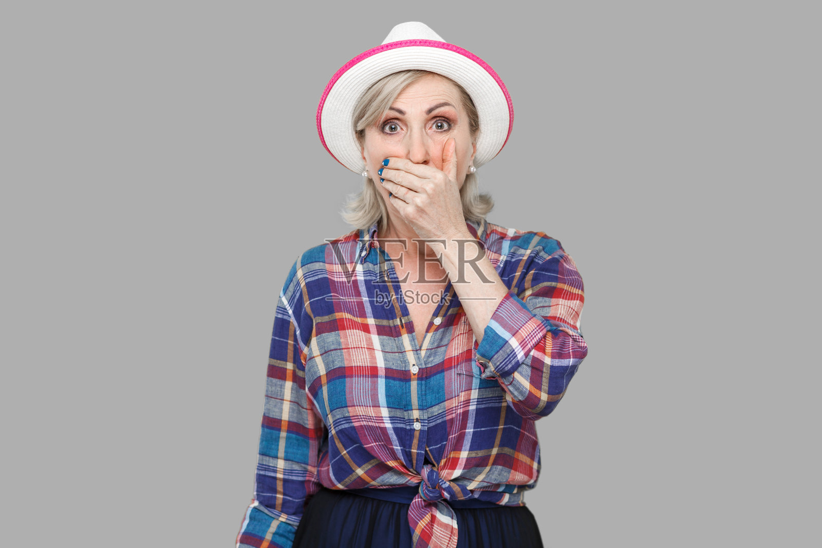 震惊时尚成熟女人的肖像在休闲风格与帽子站着，盖住她的嘴，看着相机与难以置信的面部表情照片摄影图片