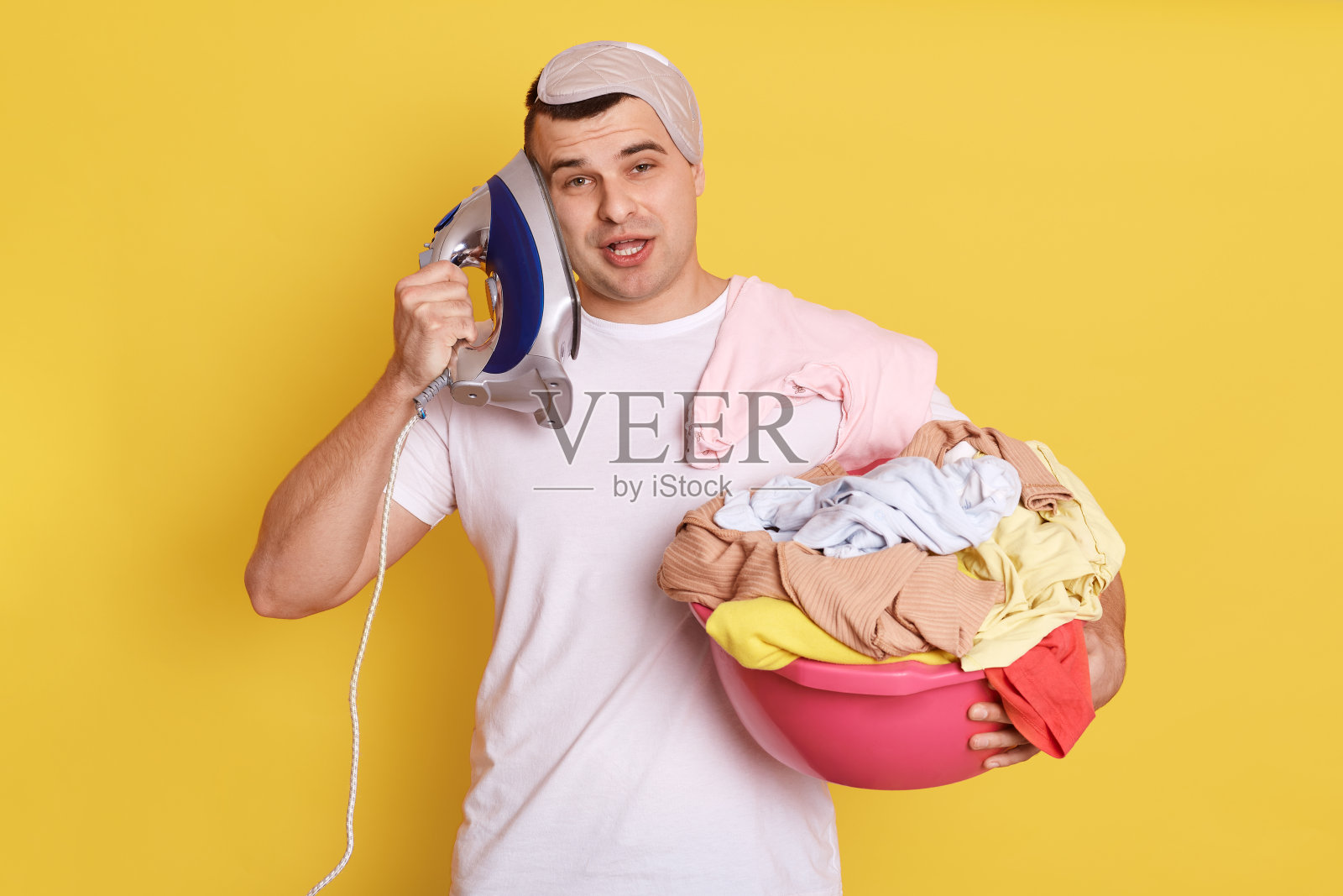 疲惫无聊的年轻男管家带着熨斗靠近耳朵，非常疲惫，想象他在打电话，需要熨烫衣服，孤立在黄色的背景下，看起来很疯狂。照片摄影图片