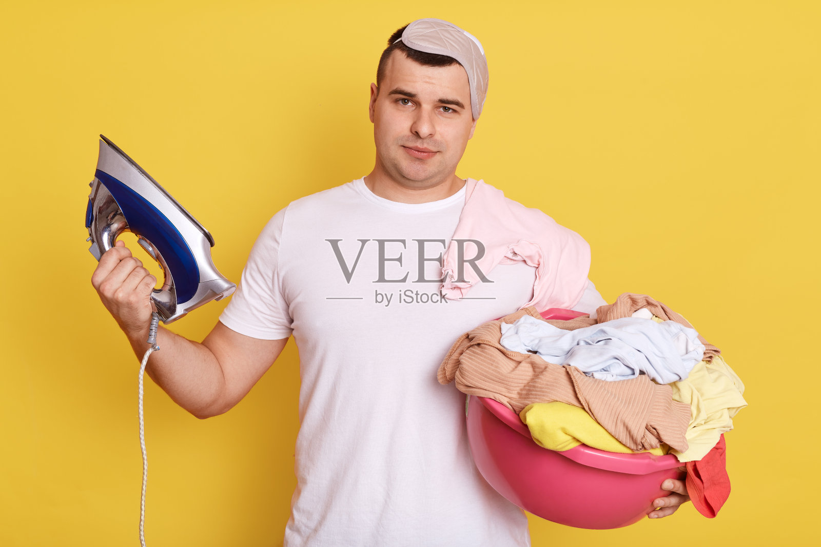 消极情绪和家务杂事，年轻的白种男人洗完衣服后，拿着盆和衣服洗，拿着熨斗，孤立在黄色的背景下，看起来疲惫不堪。照片摄影图片